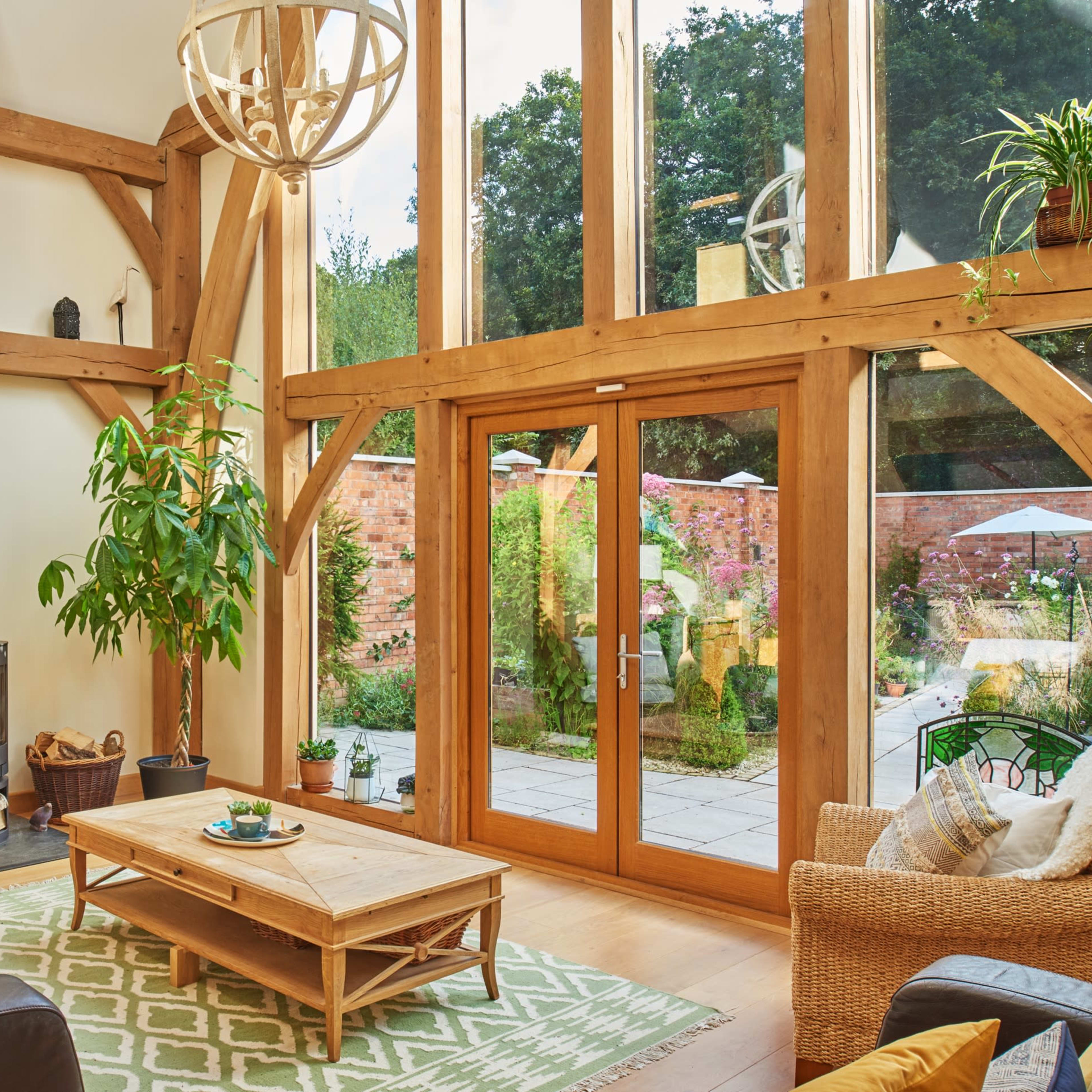 The scene features a bright living room with wooden beams, a wood-burning stove, a large glass door opening to a garden, and a table in the center surrounded by various seating.