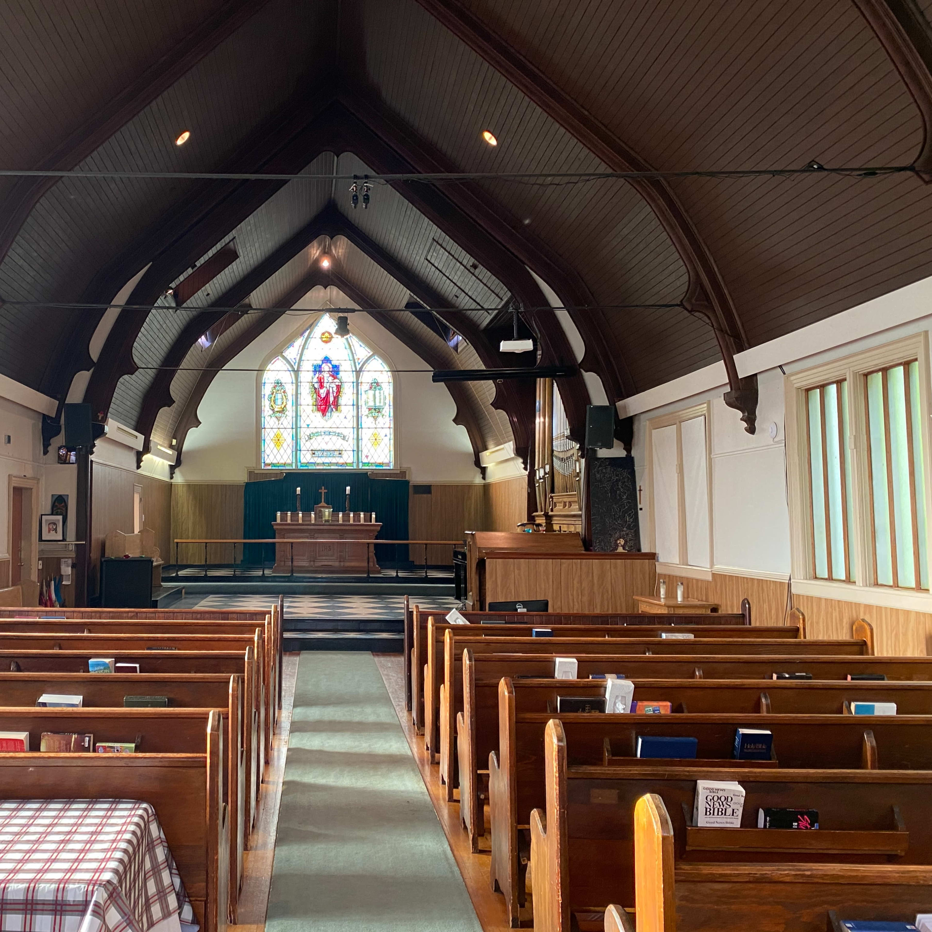 The interior of a church features a row of wooden pews facing an altar with a stained glass window in the background.