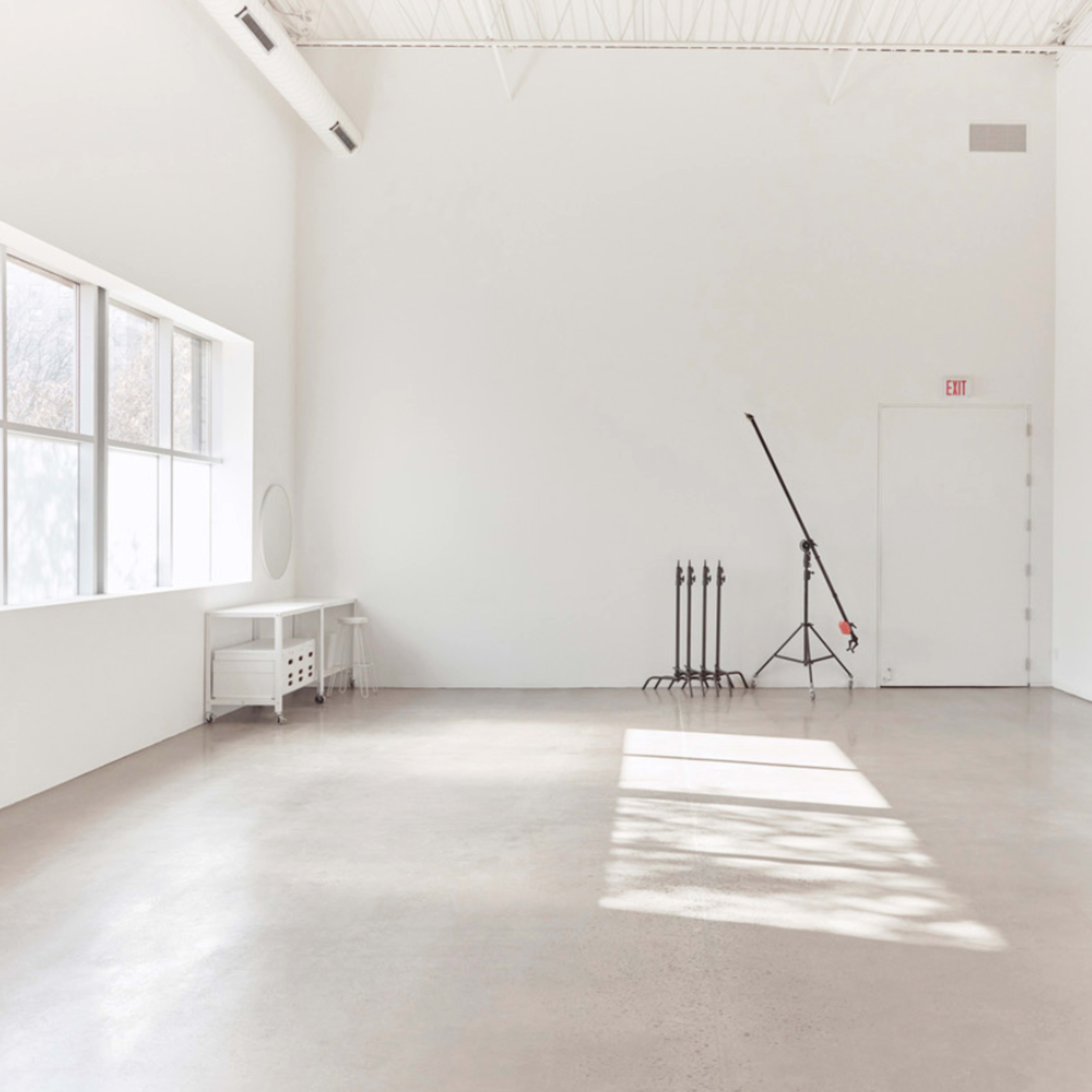 A bright, empty room with white walls, a large window, a door, and a lighting stand in the corner.