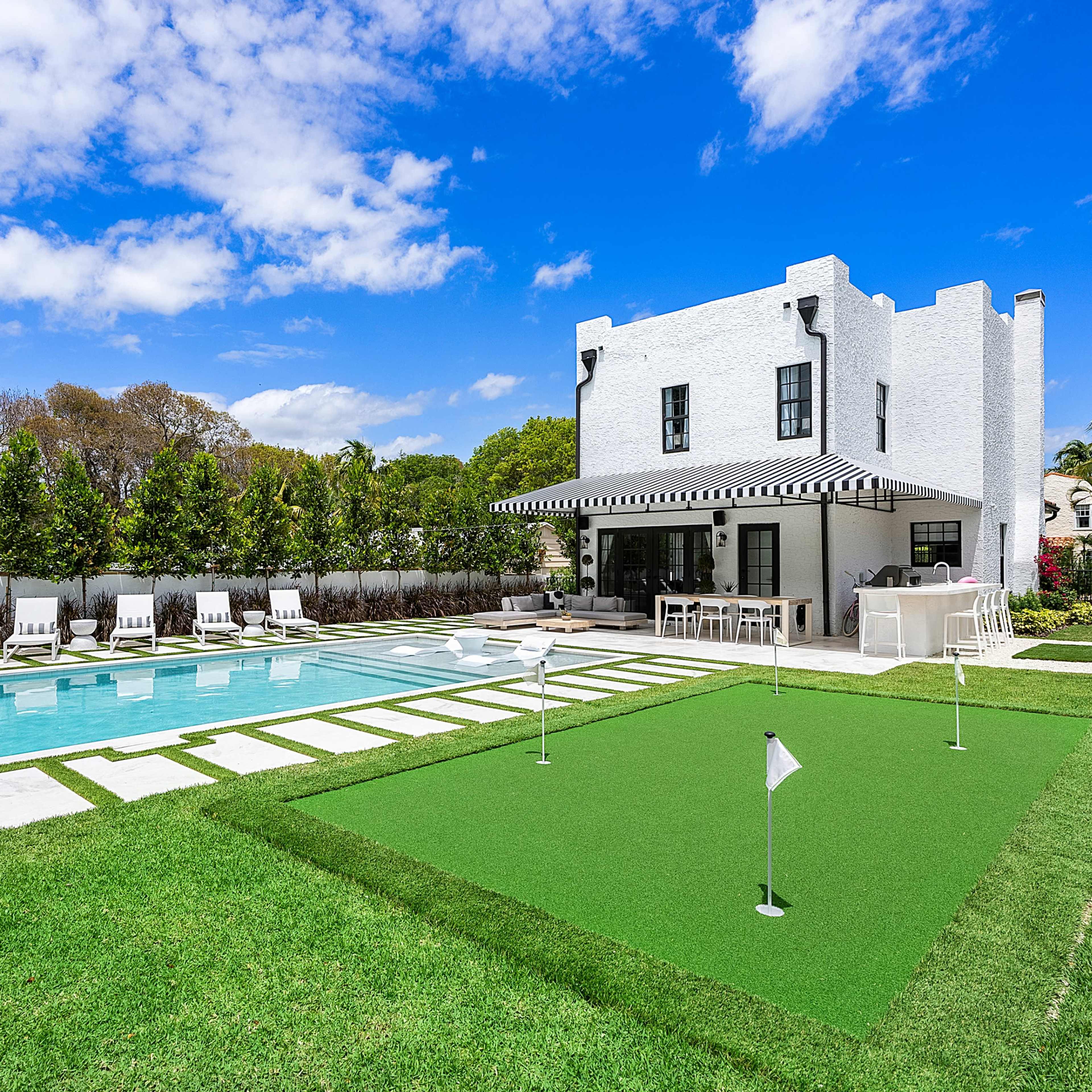 A modern white house with a pool and putting green surrounded by manicured lawns and shrubs.