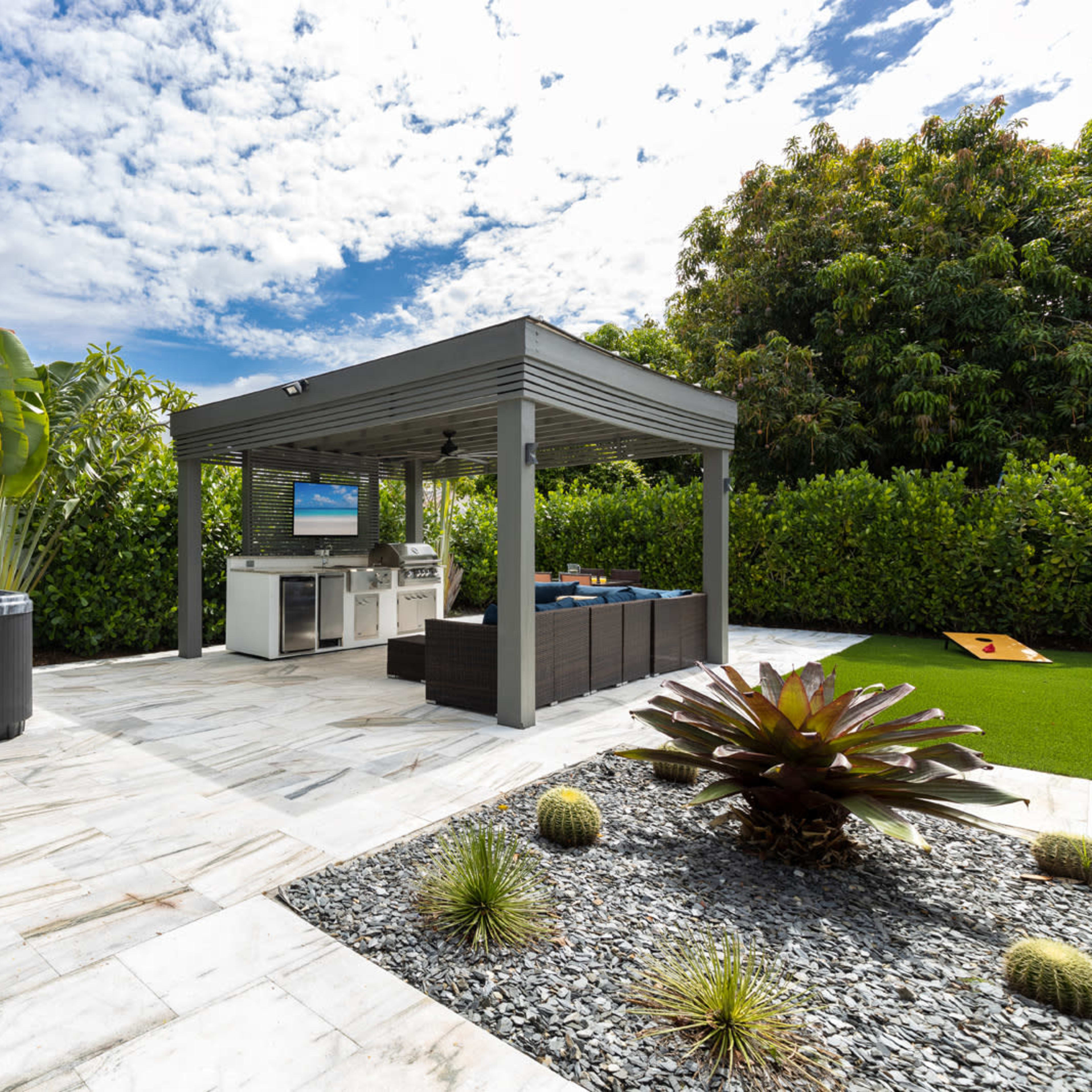 A landscaped outdoor area featuring a hot tub, a covered cooking space with a television, and a neatly arranged garden with stone pathways and plants.