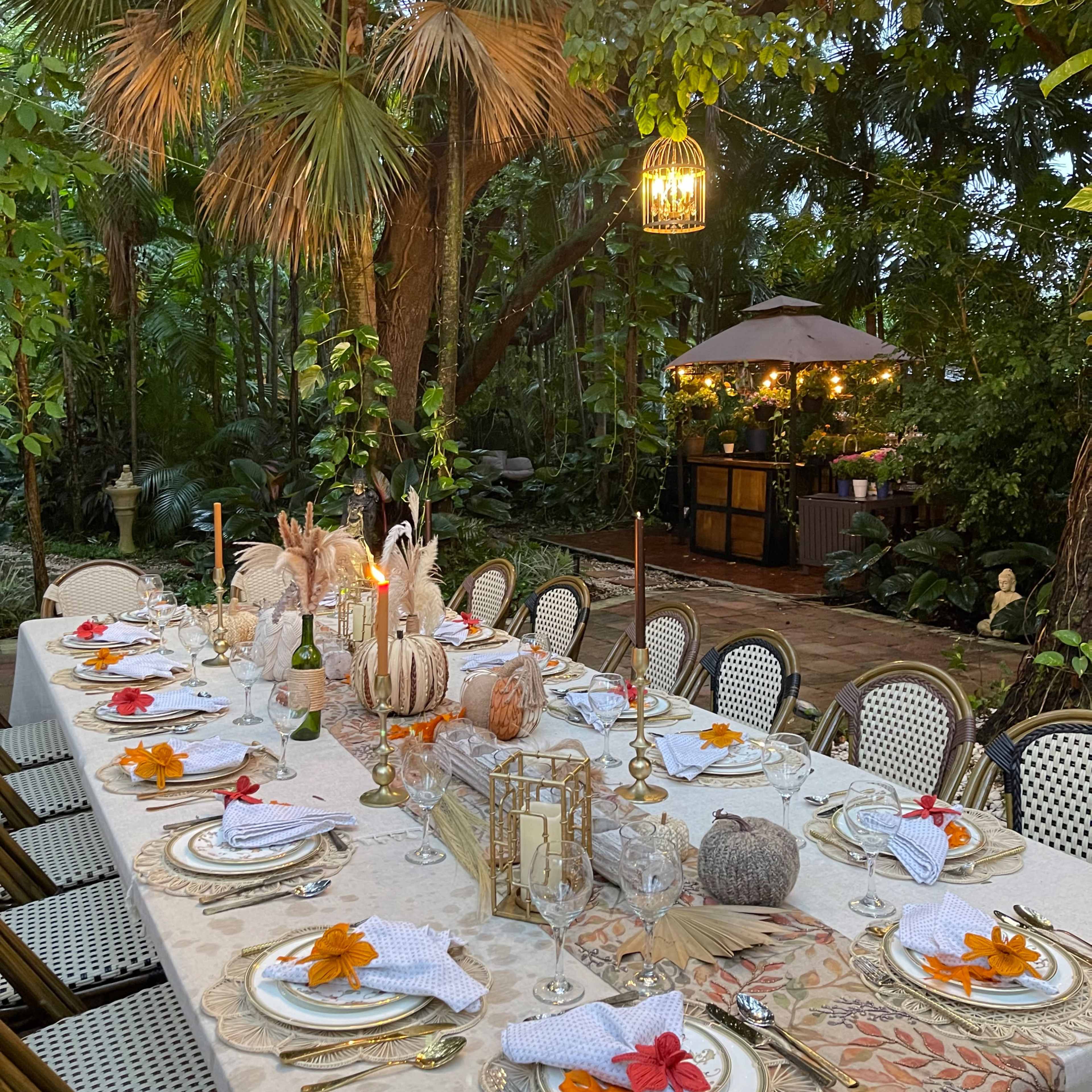 A long dining table is set with elegant tableware and decorations in a lush, green outdoor setting illuminated by a chandelier and soft lighting.