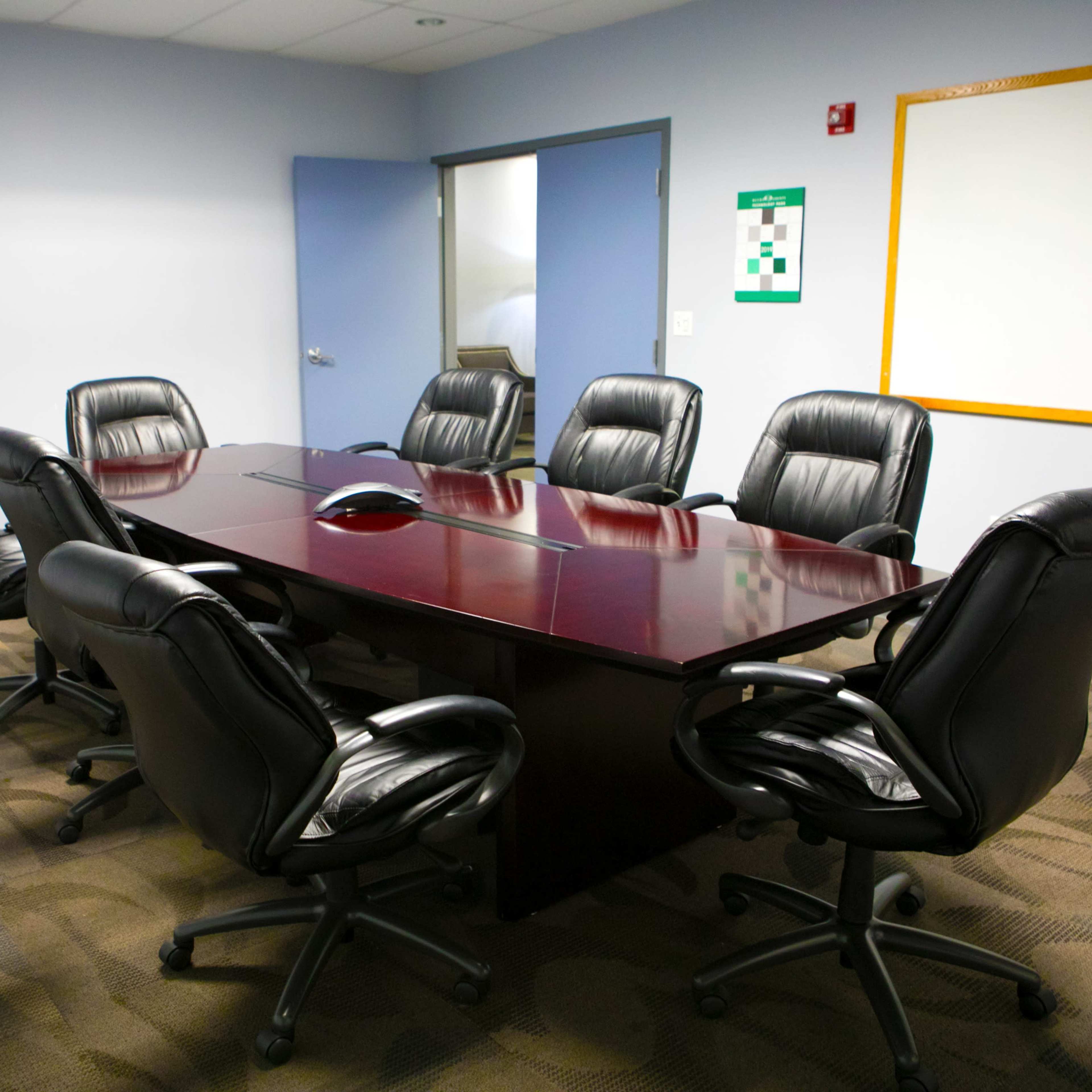 A conference room features a large wooden table surrounded by six black ergonomic chairs, with a whiteboard and emergency exit sign on the walls.