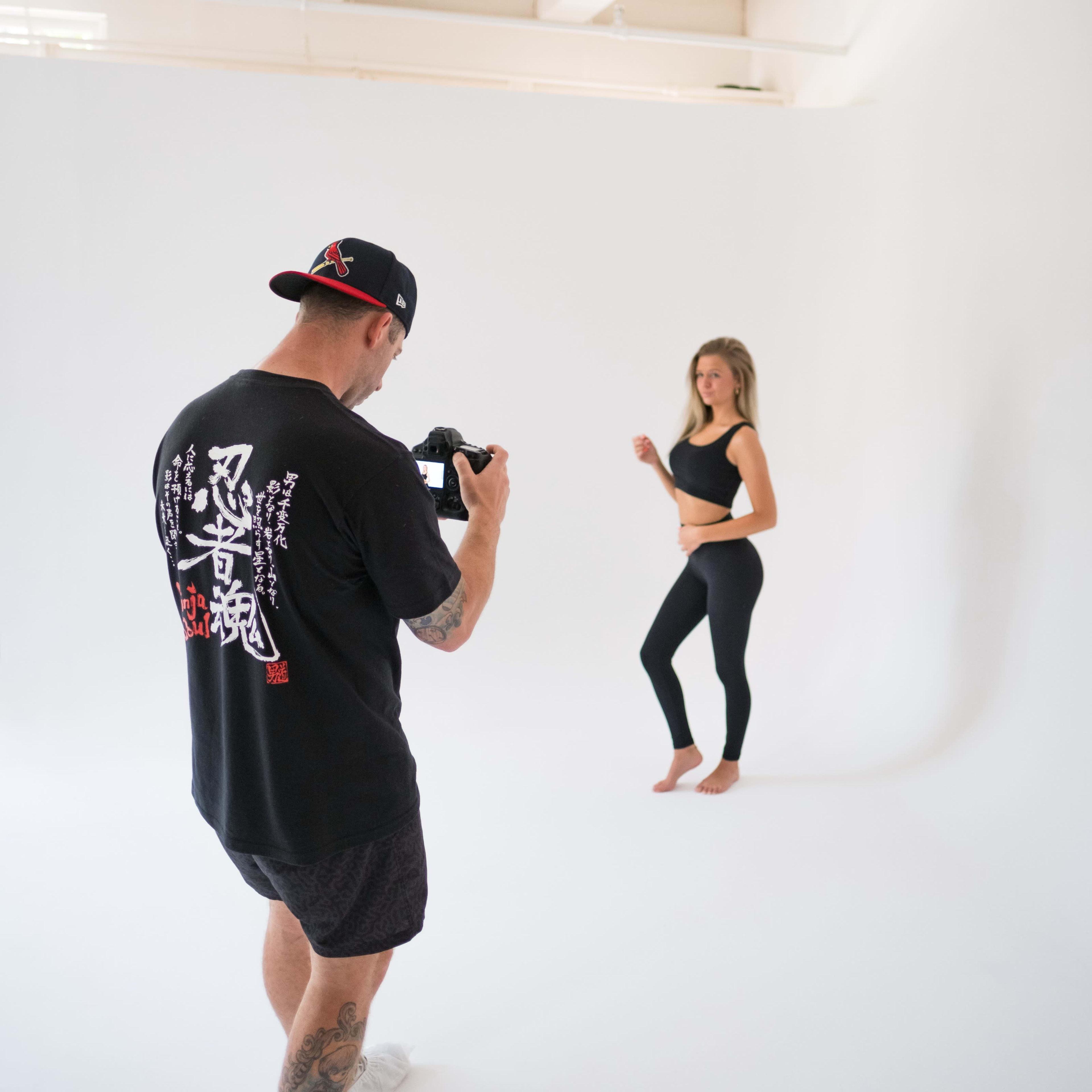 A photographer is taking pictures of a model in a studio with a white backdrop.