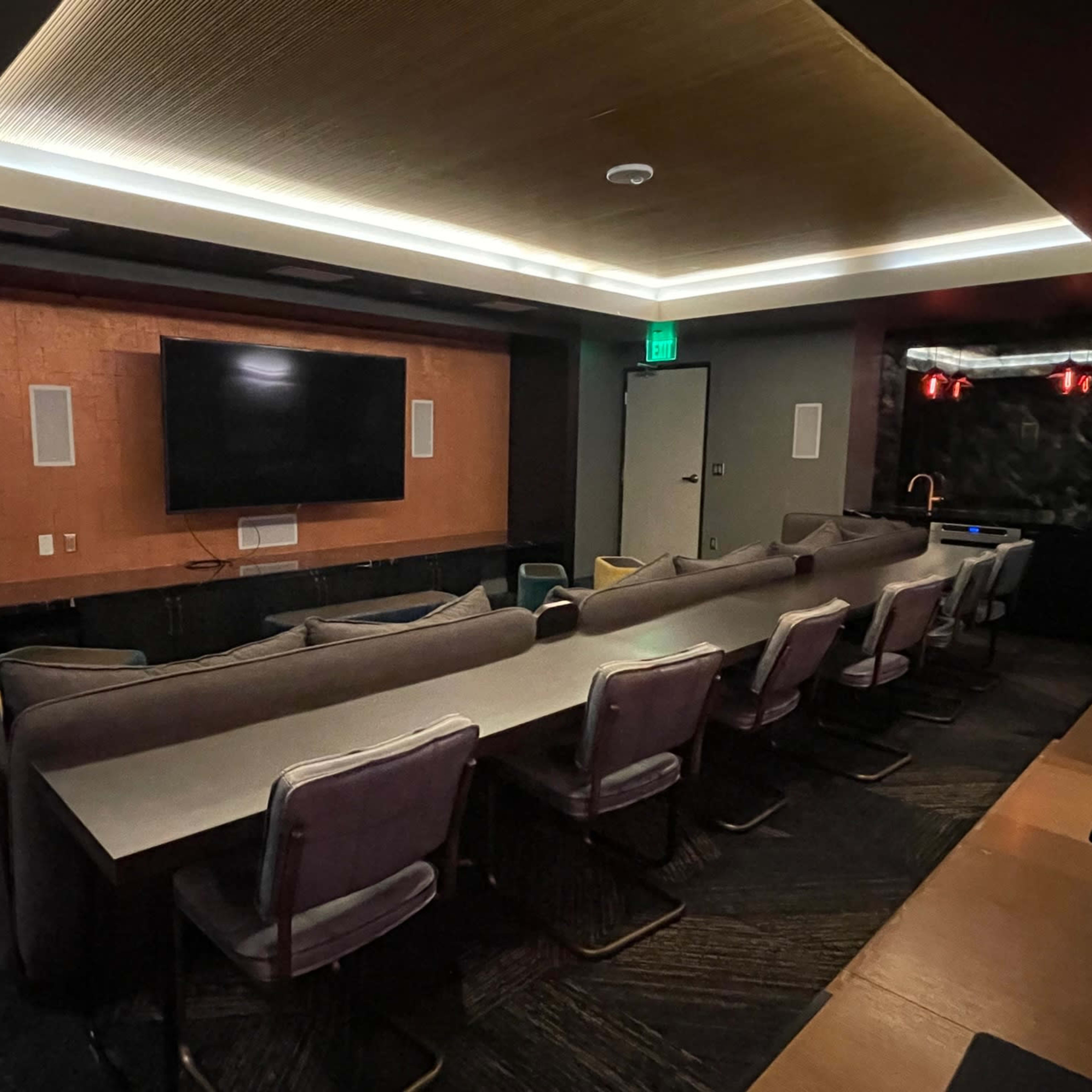 A modern, dimly lit room with a long table surrounded by chairs, a large wall-mounted television, and a textured wall panel behind the seating area.