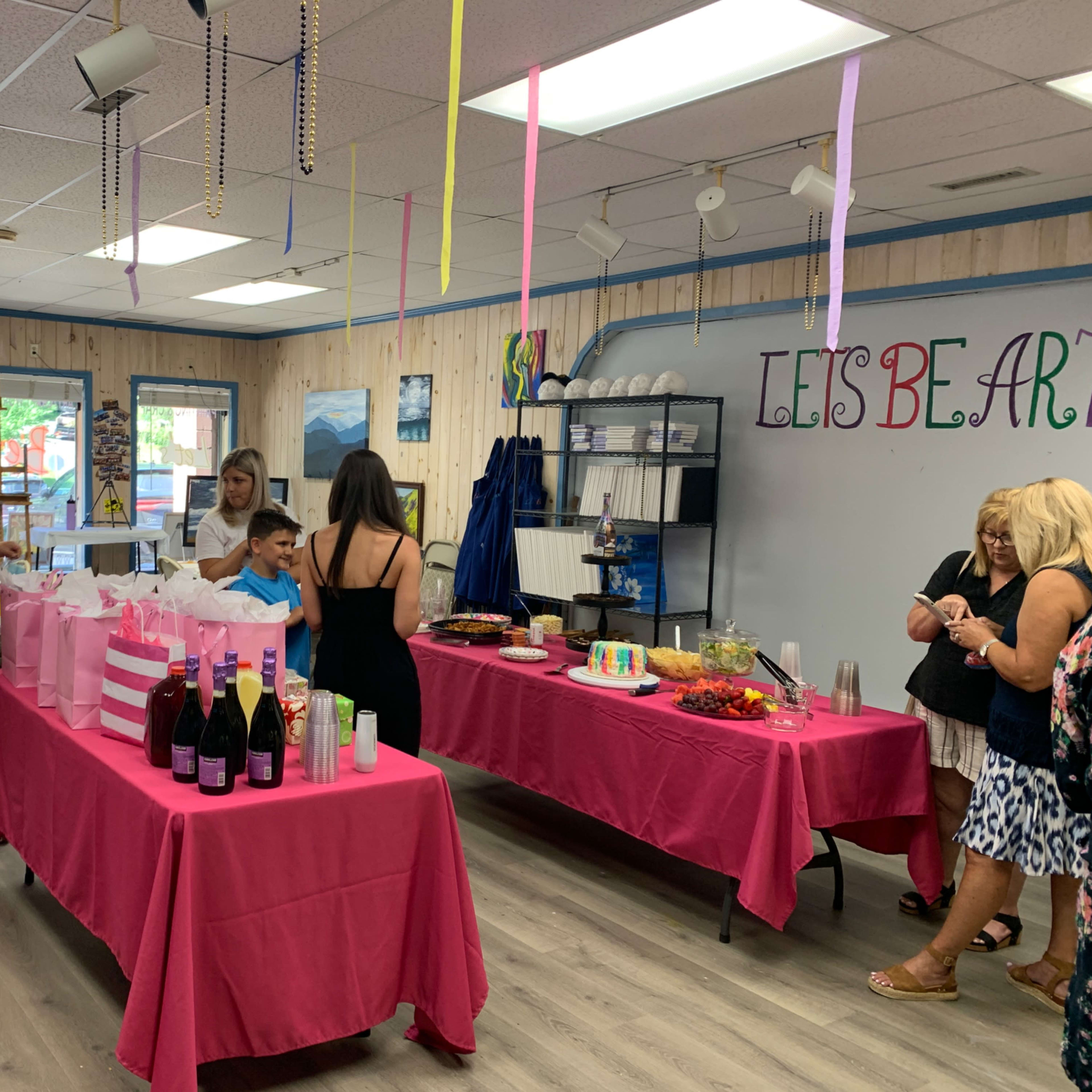 A celebration venue with guests mingling around pink tables adorned with food and drinks.