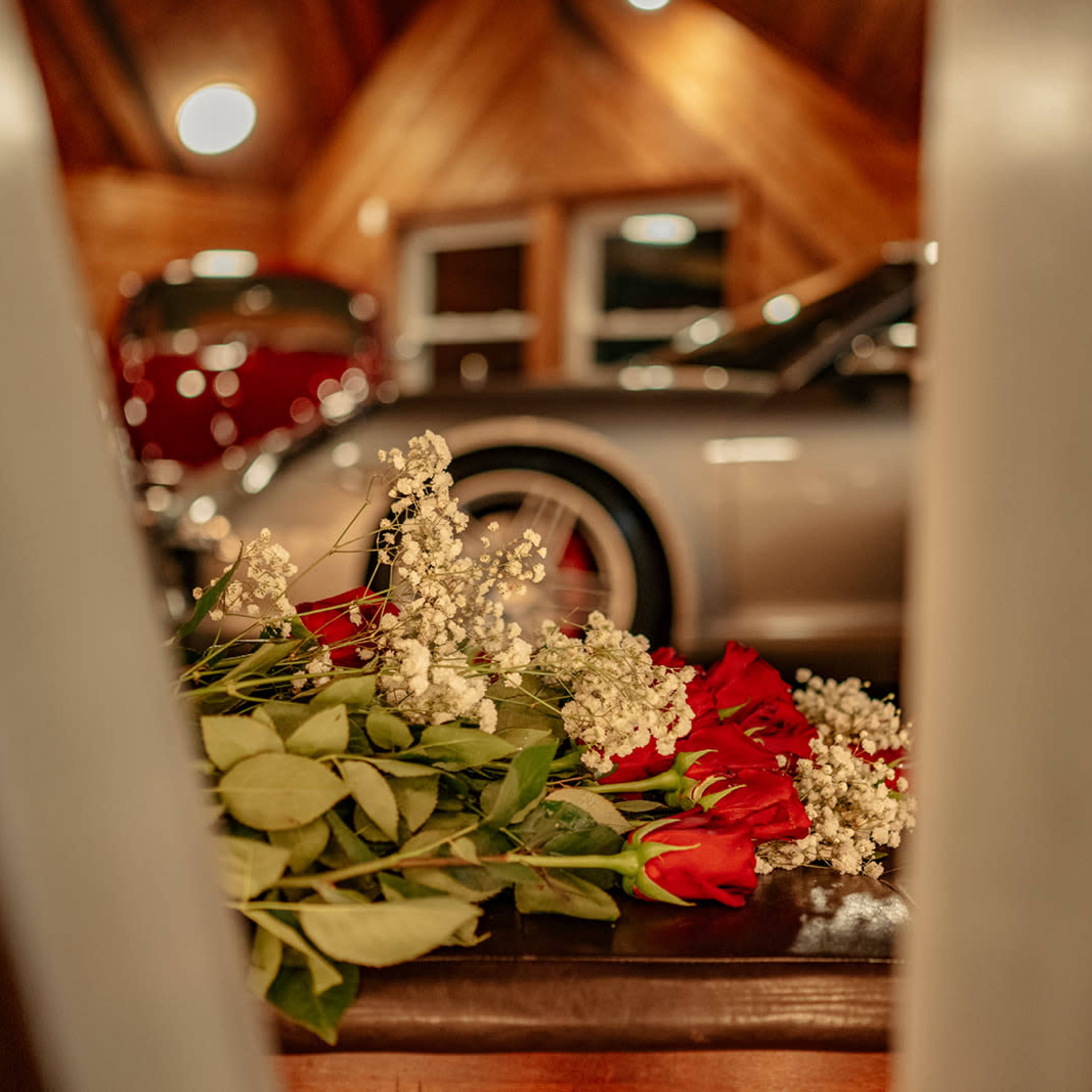 A bouquet of red roses and white flowers rests on a table near vintage cars parked in a wooden-paneled garage.