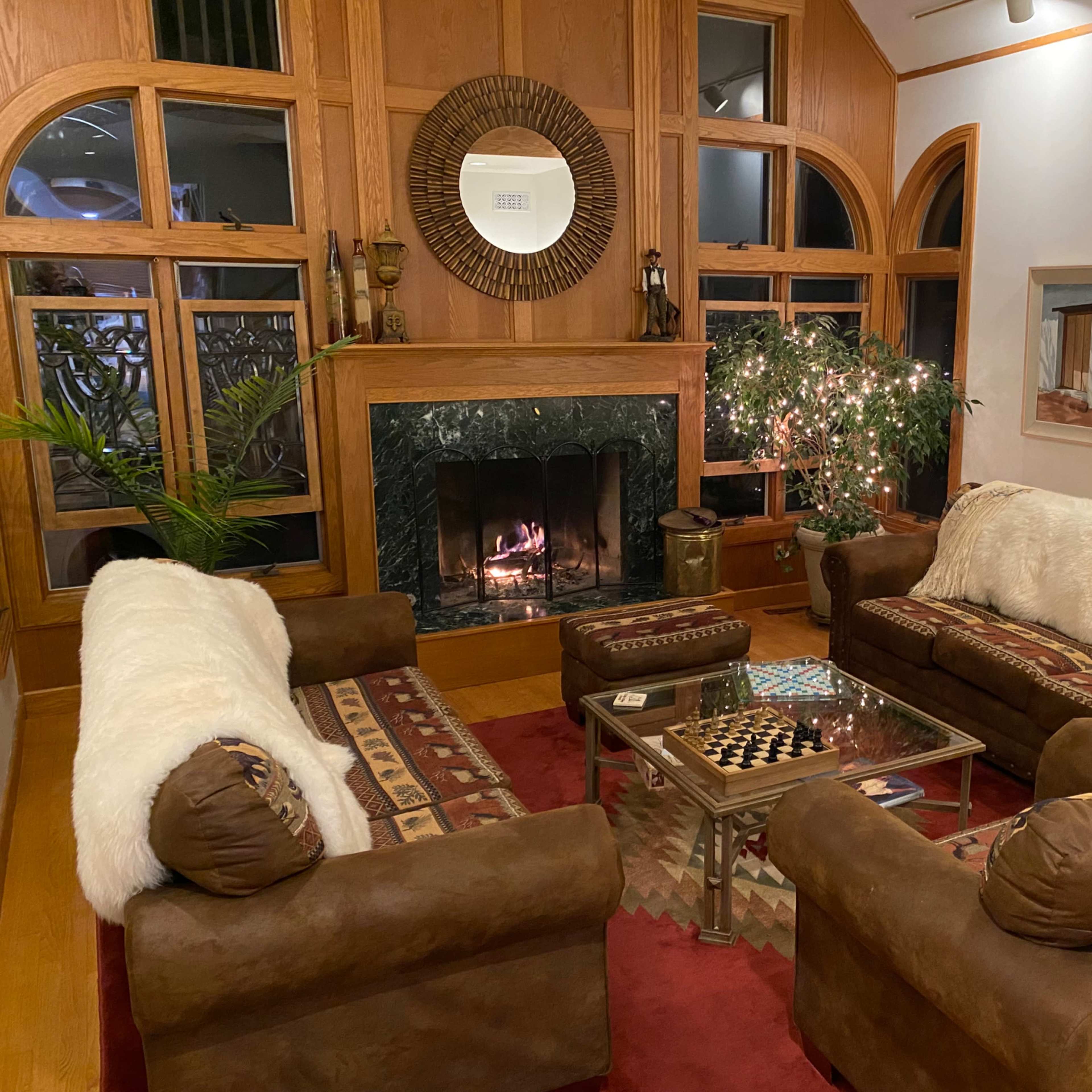 The scene features a cozy living room with two brown sofas, a glass coffee table, a warm fireplace, and a decorative plant surrounded by various artworks on the walls.