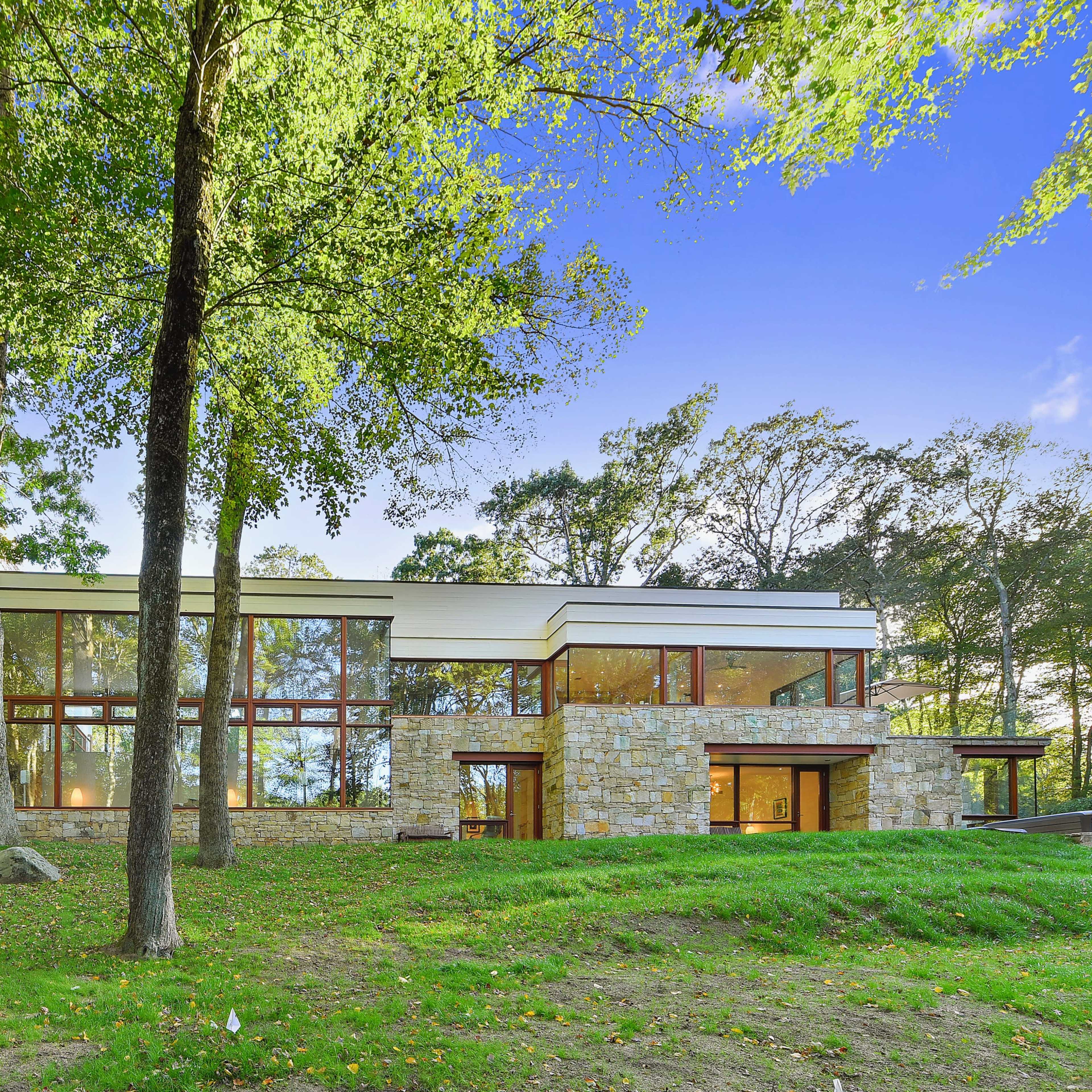 A modern house with large glass windows and stone accents, set in a green landscape with trees.