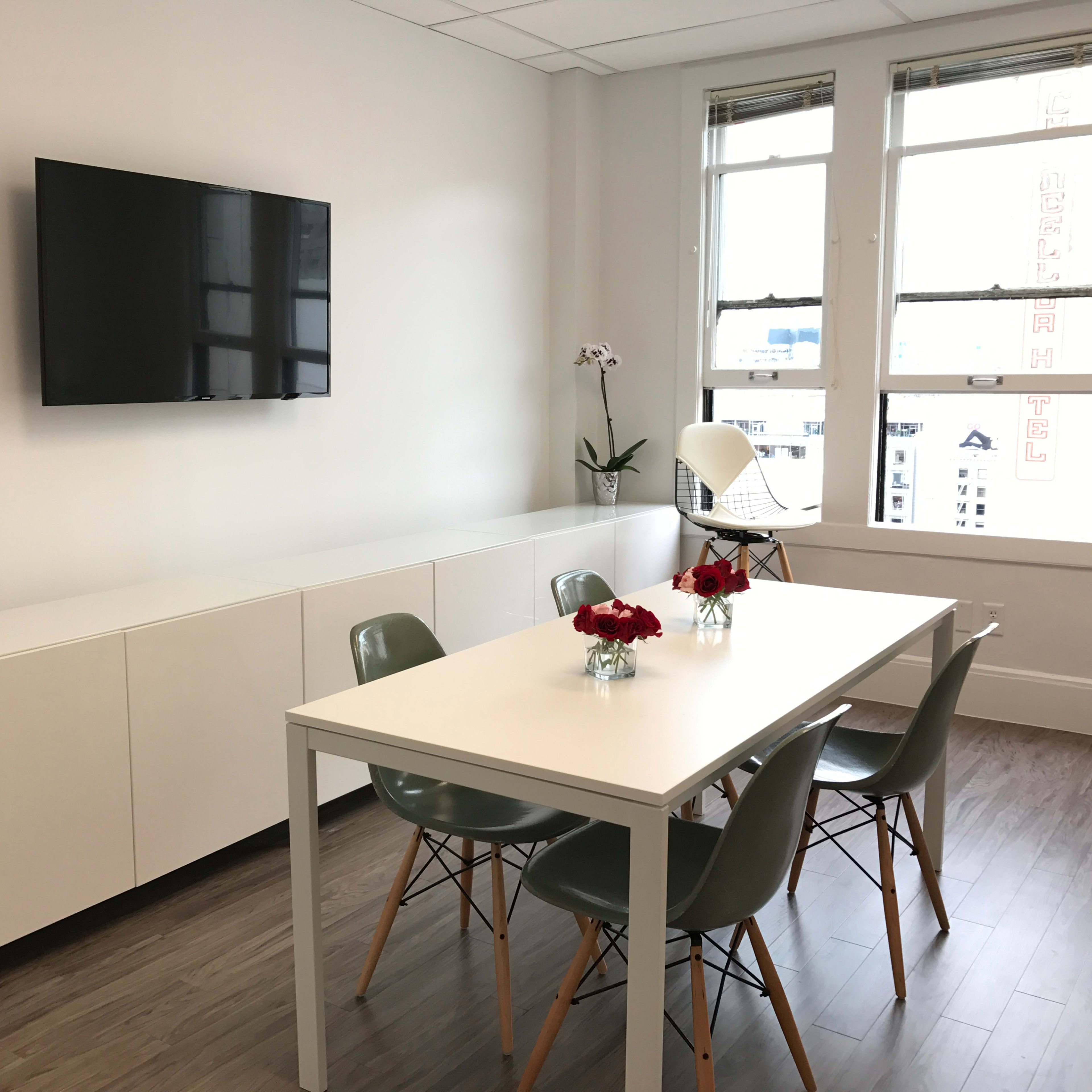 A modern conference room features a white table with four chairs, a television mounted on the wall, and large windows letting in natural light.