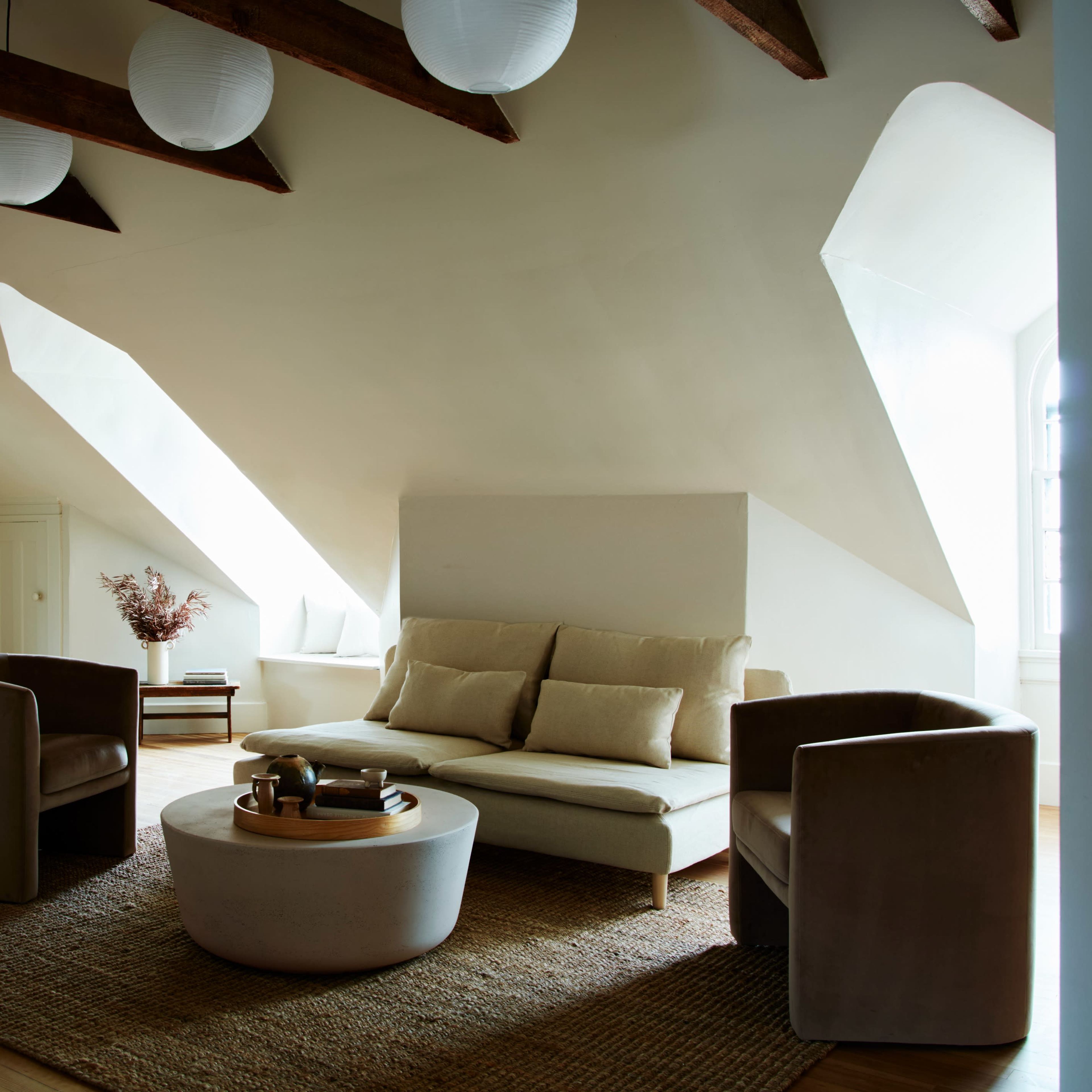 A spacious living room features a modern sofa, two armchairs, and a round coffee table, all set against slanted ceilings and large windows.