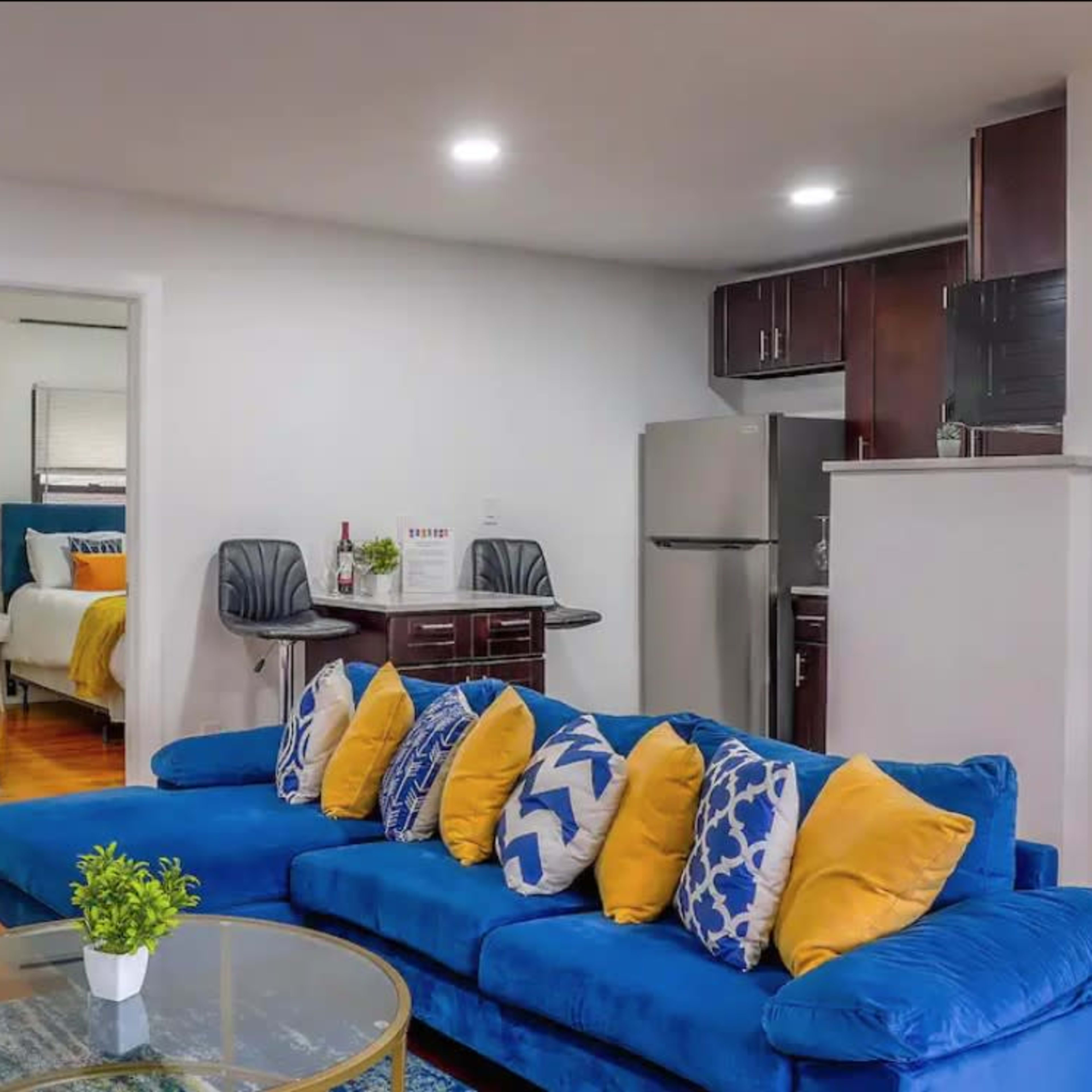 A modern living room with a blue sofa adorned with yellow and white pillows, a glass coffee table, and a view into a kitchen and adjacent bedrooms.