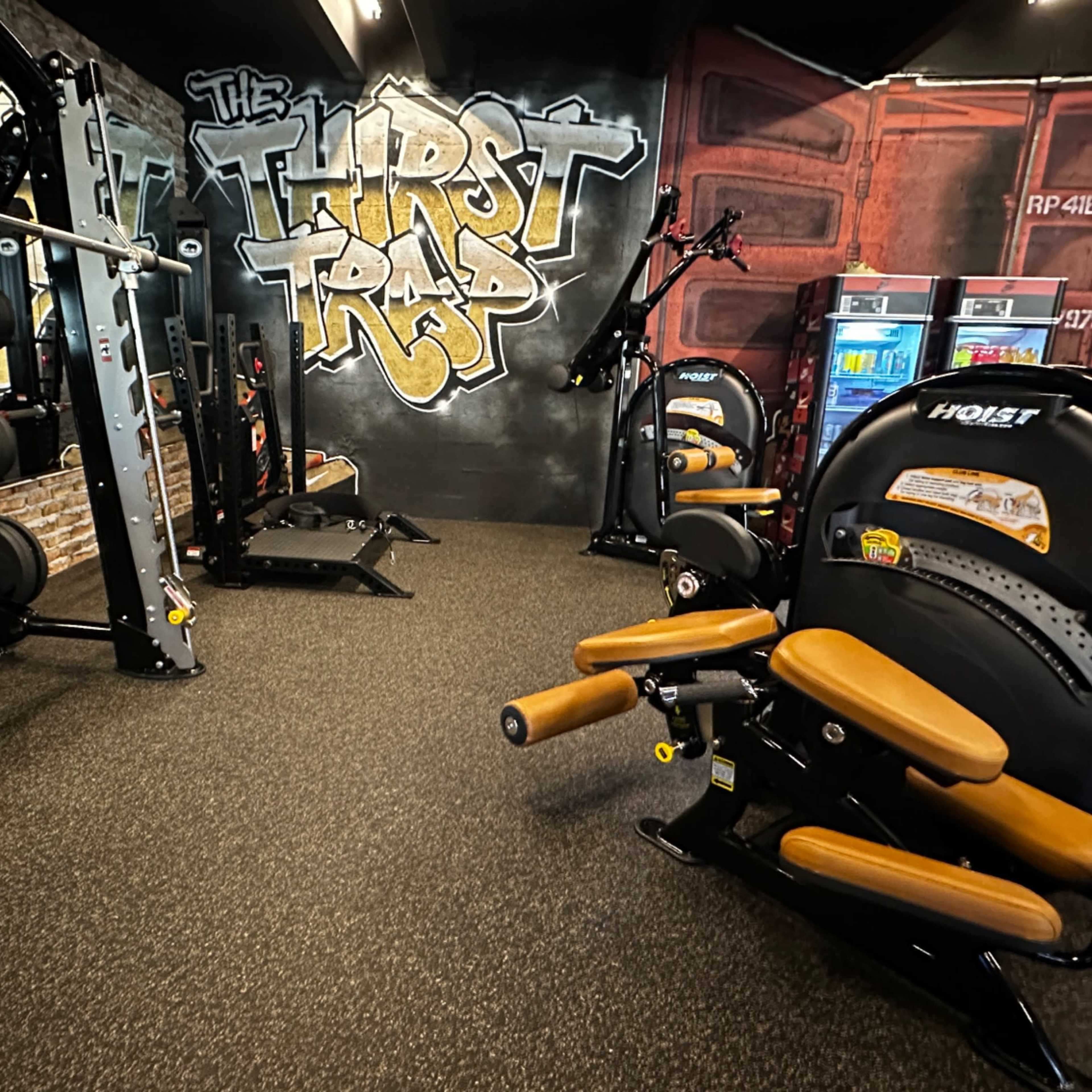The gym features a variety of strength training equipment and a graffiti-style mural on the wall, labeled "The Thirst Trap."