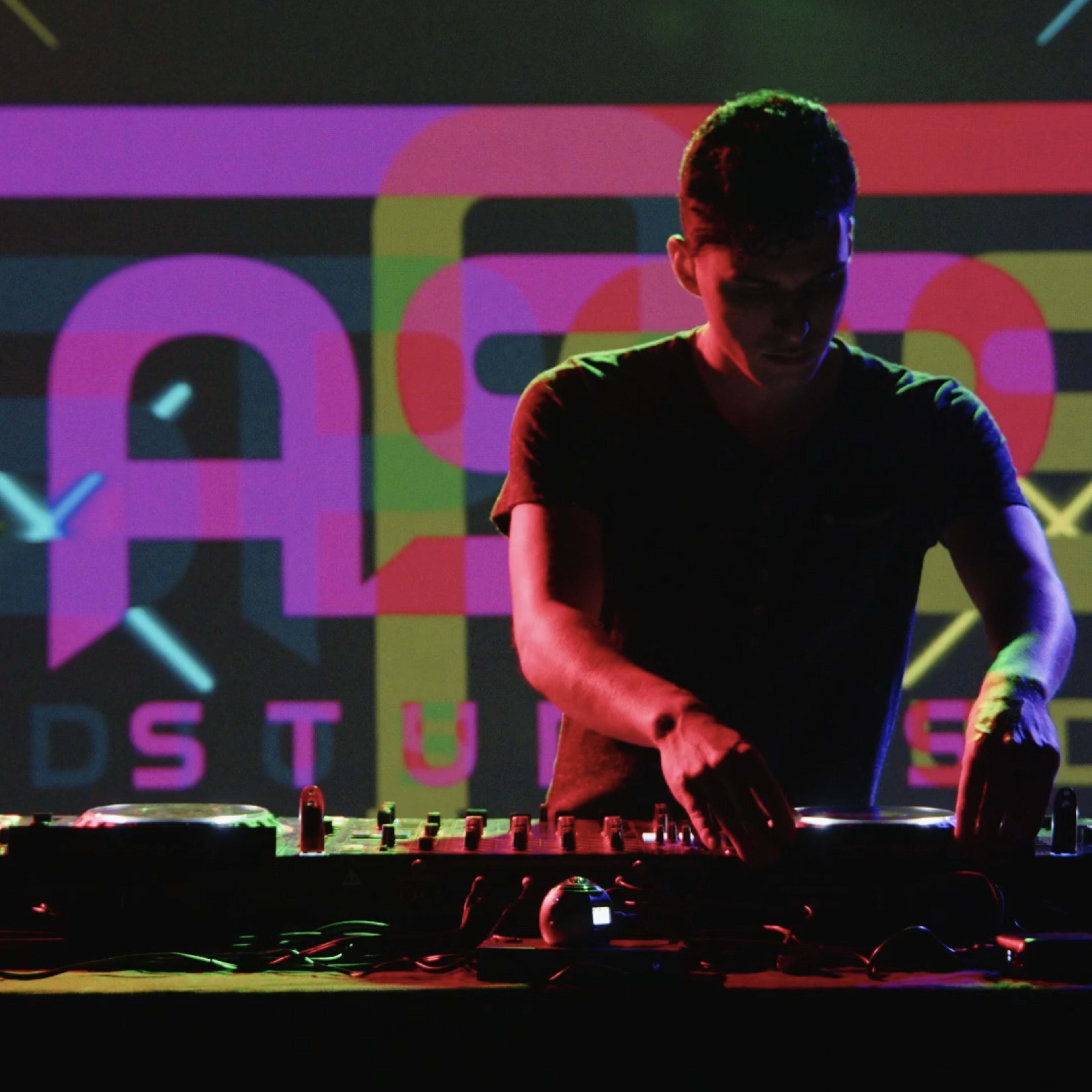 A DJ manipulates a mixing console in front of a brightly lit backdrop featuring colorful graphic elements.