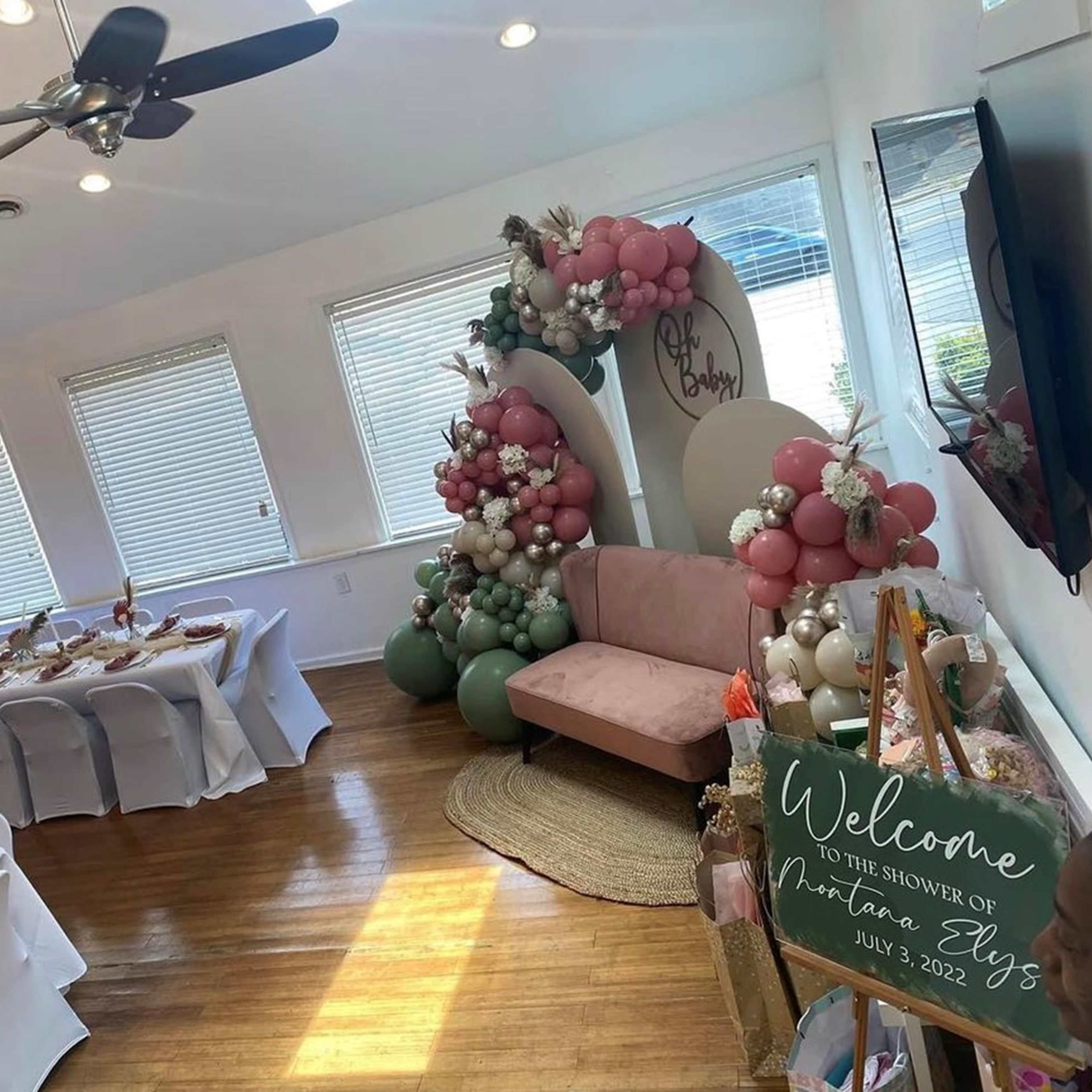 A decorated room set up for a baby shower, featuring a long dining table, chairs covered in white fabric, and a seating area adorned with balloons and a welcome sign.