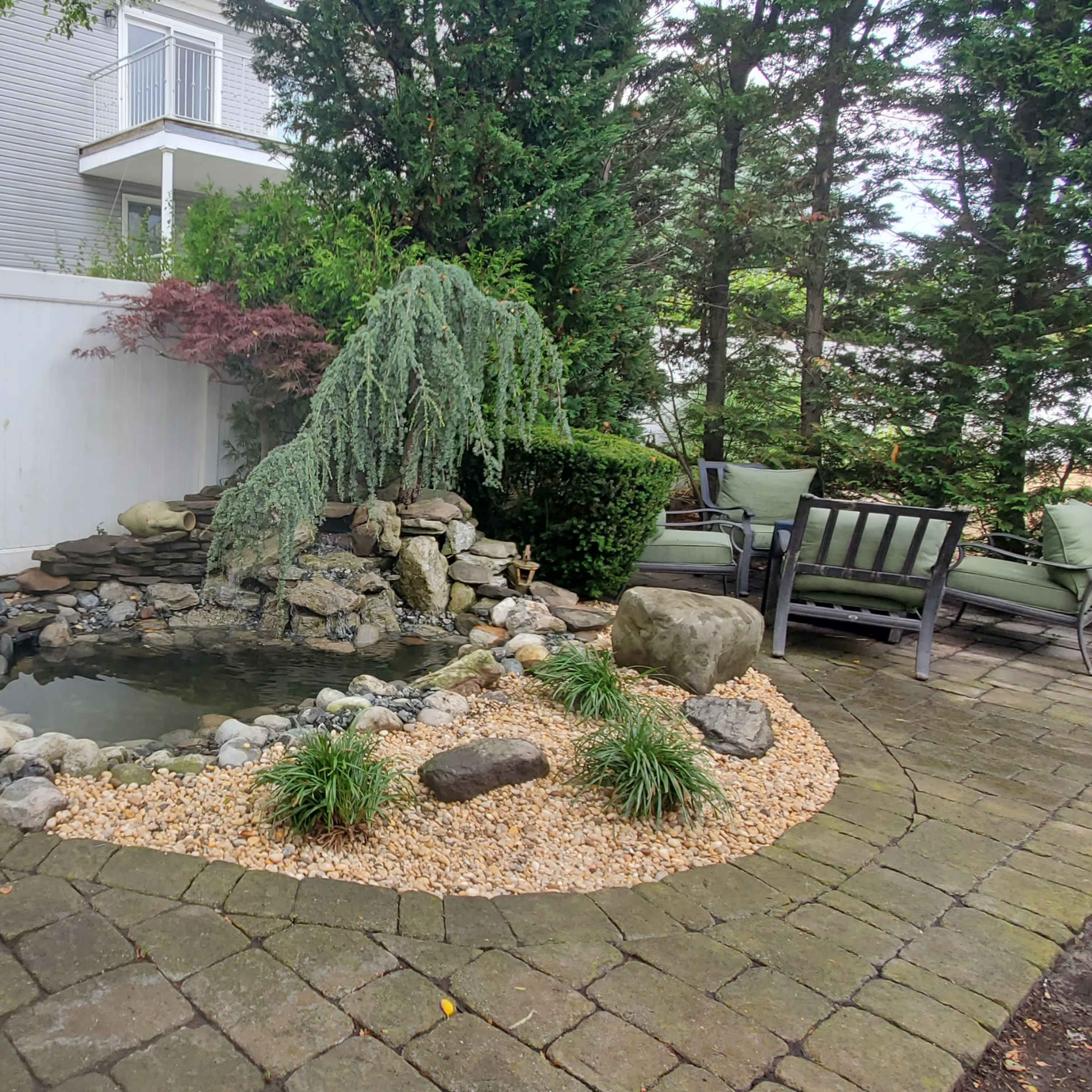 A landscaped garden featuring a small pond surrounded by rocks, gravel, and various green plants, with a seating area visible nearby.