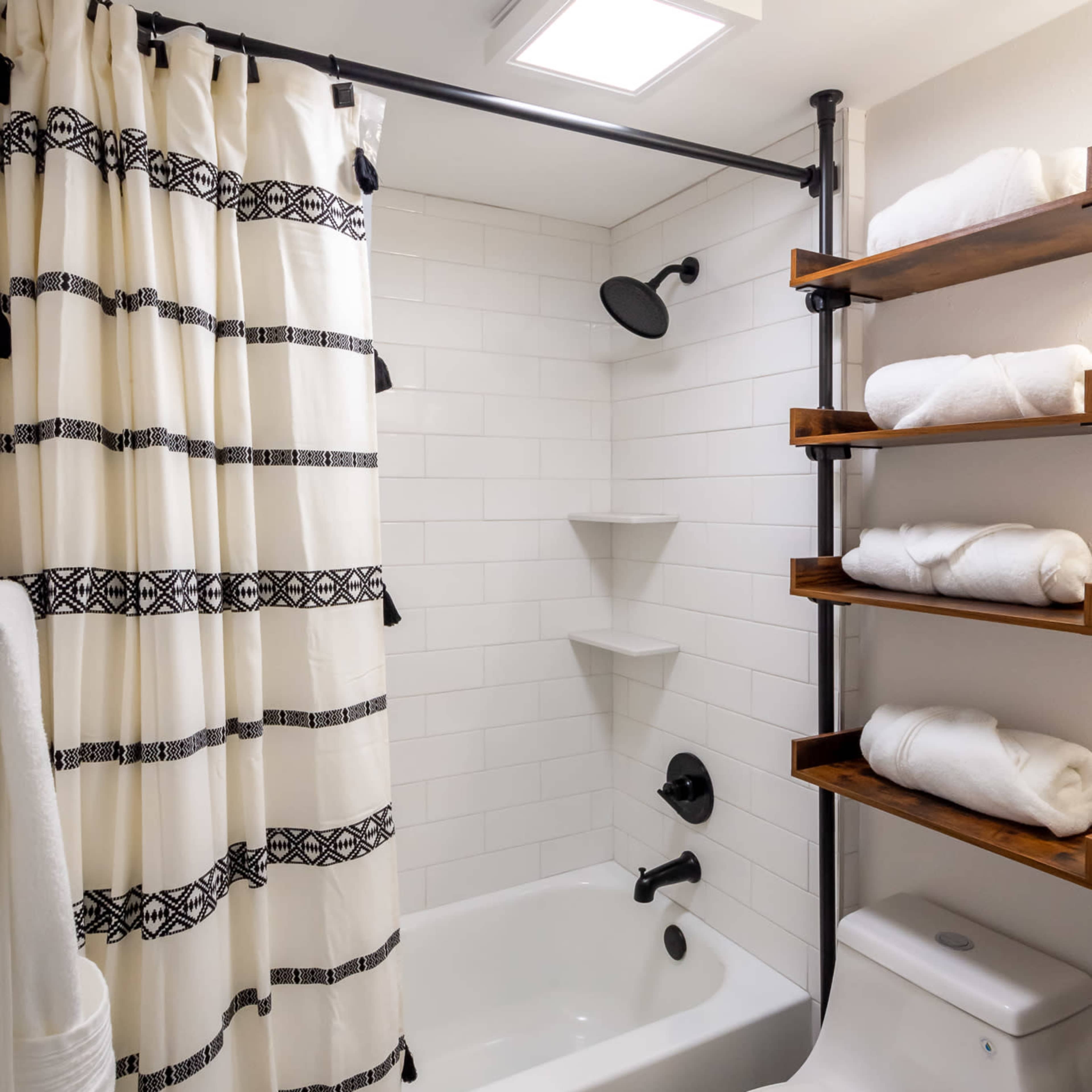 A bathroom featuring a bathtub with a shower curtain, a showerhead, a toilet, and wooden shelves holding neatly stacked white towels.