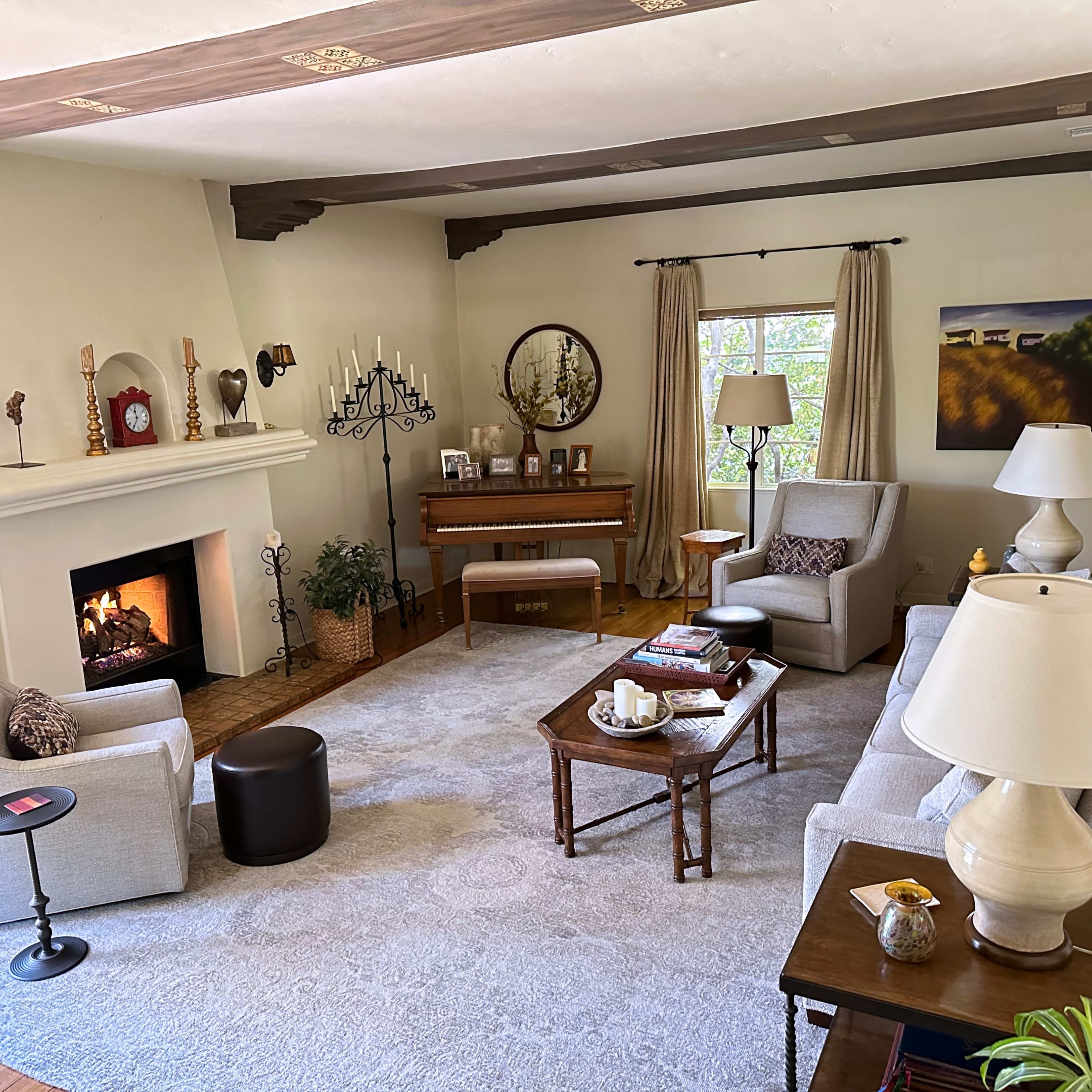 A cozy living room featuring a fireplace, a piano, two armchairs, and a sofa arranged around a large area rug.