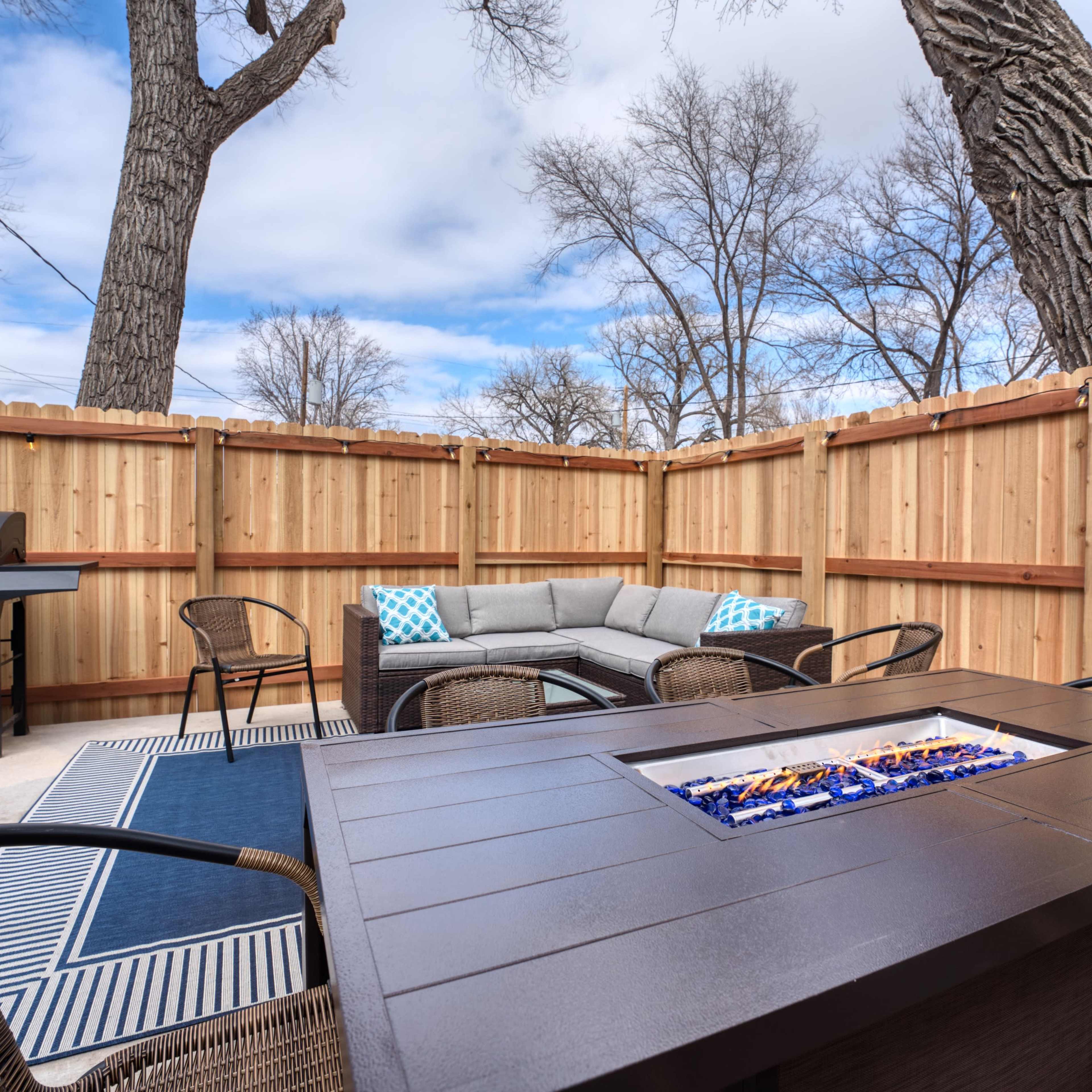 A cozy outdoor patio area with a fire pit, seating arrangement, and a barbecue grill surrounded by wooden fences and trees.