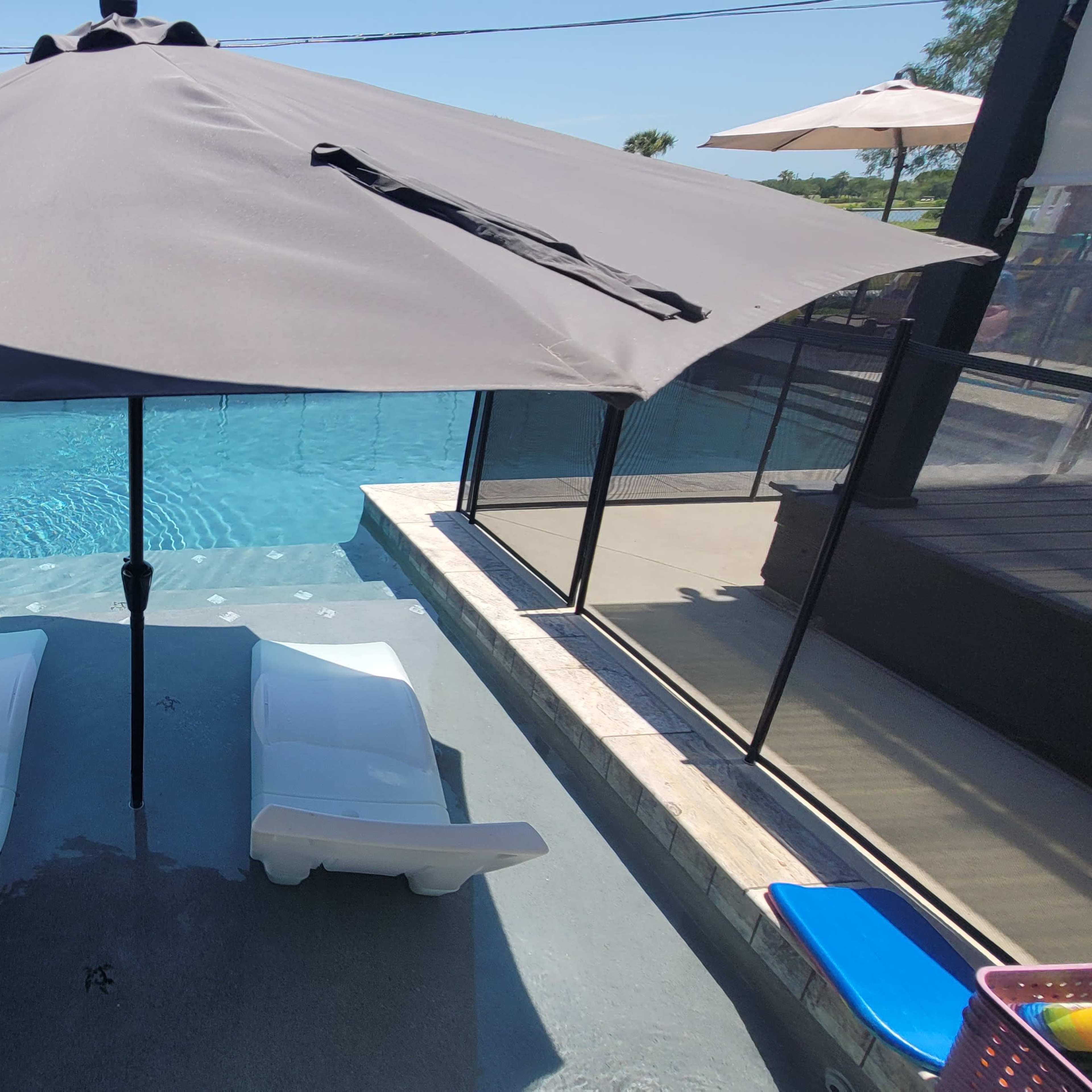 A pool area with two white lounge chairs beneath a large umbrella, surrounded by a safety fence and water.