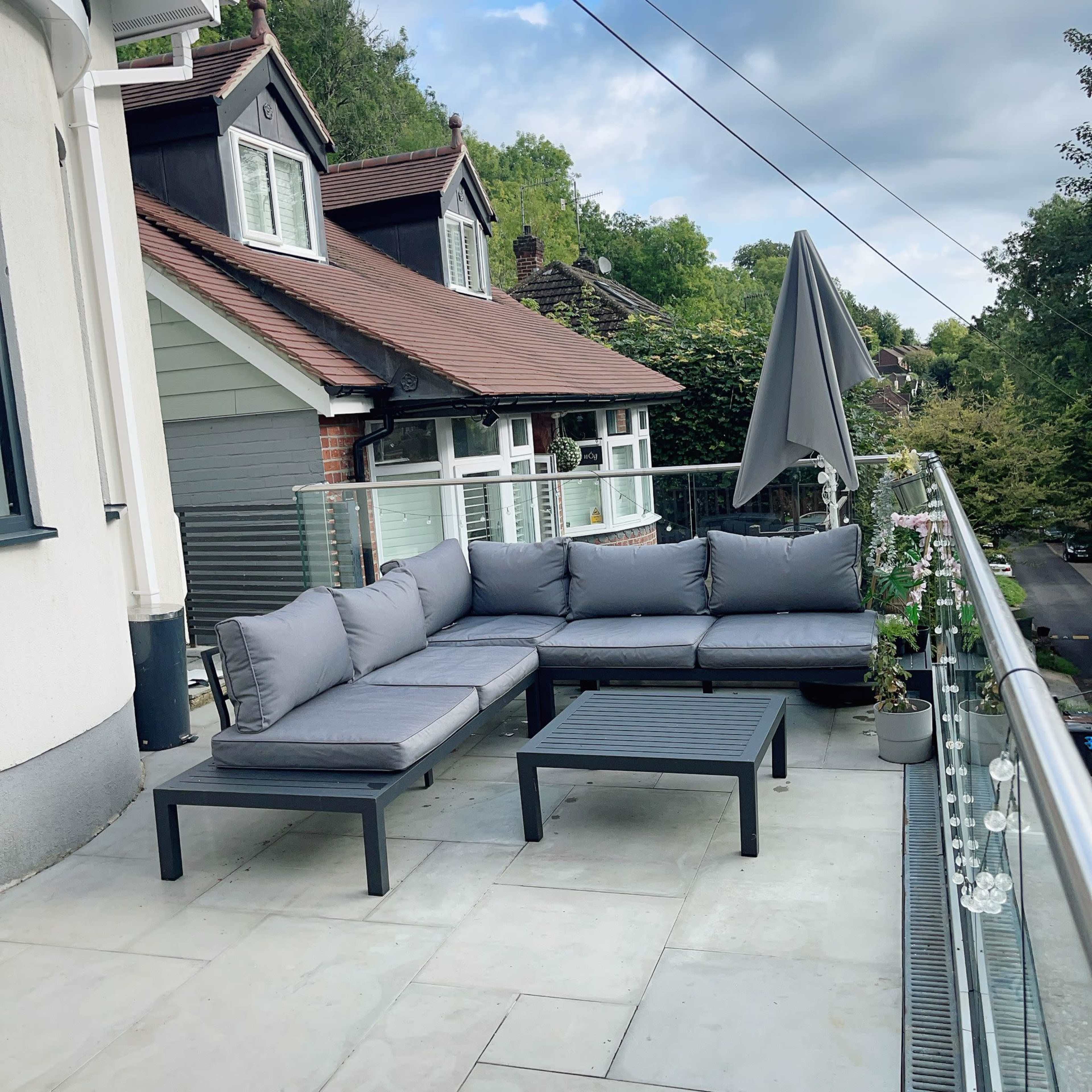 A modern outdoor seating area features a gray sectional sofa and a small coffee table on a balcony with a view of nearby houses and trees.
