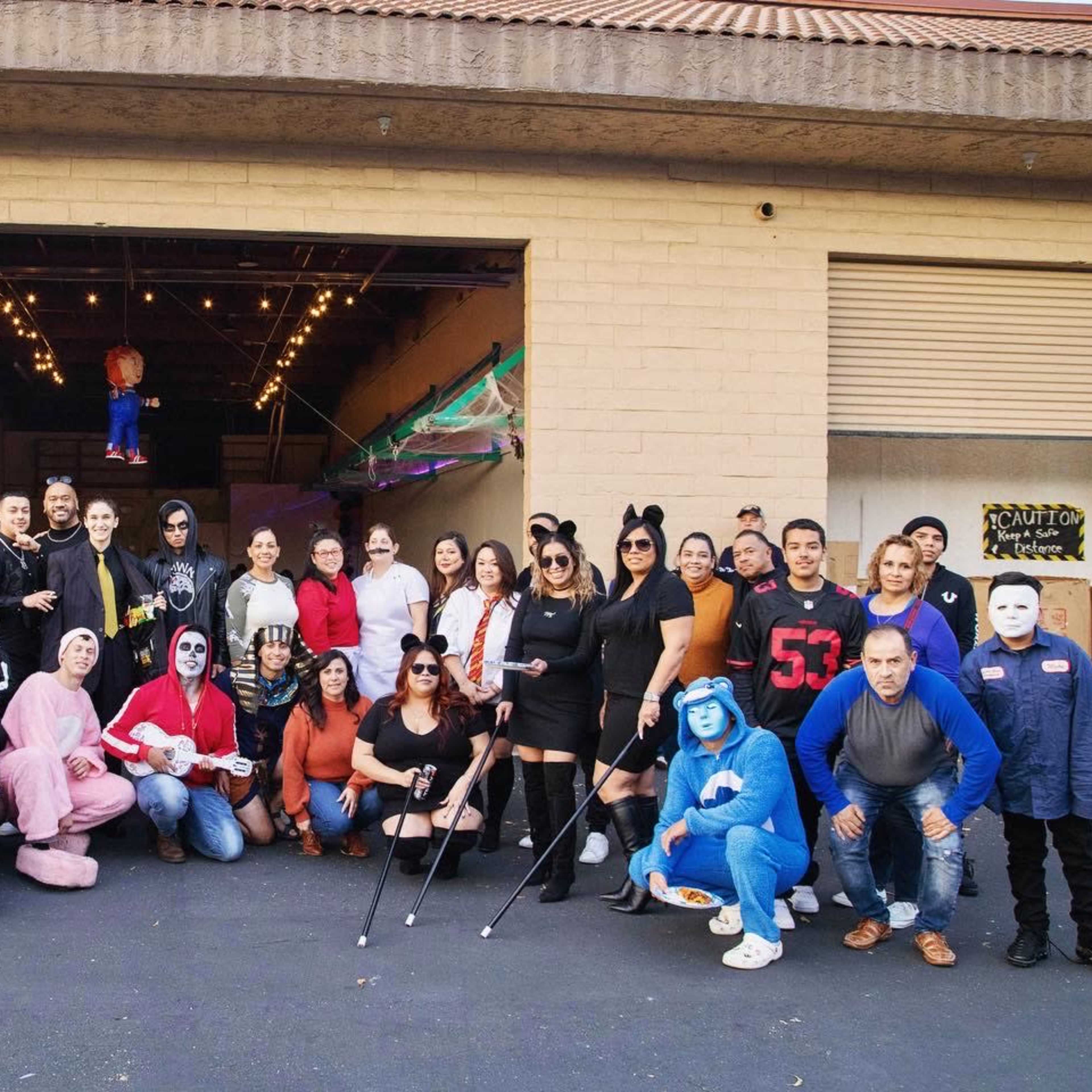 A large group of people poses together in front of a building, many dressed in various costumes and masks for a themed event.