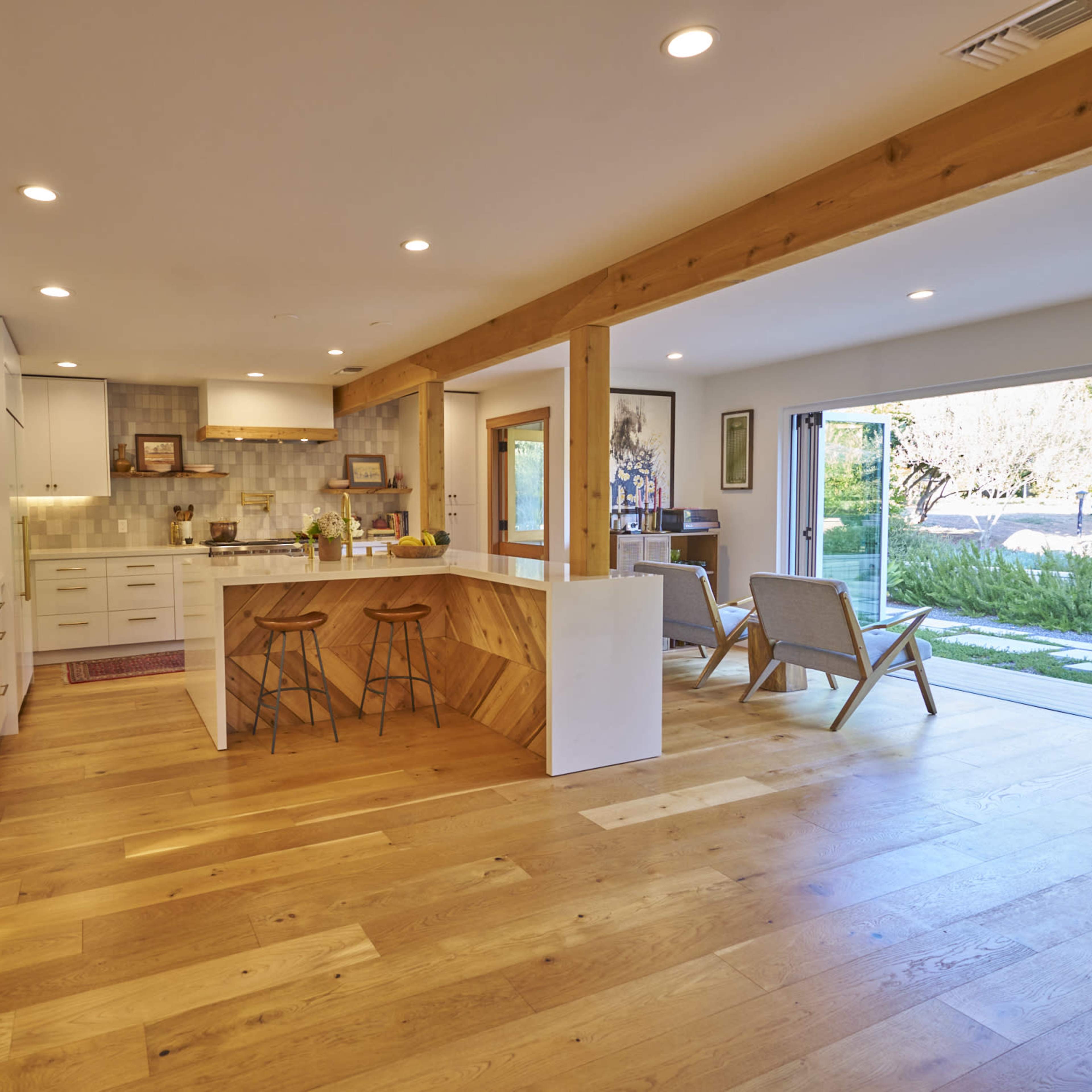A modern kitchen features a central island with bar stools, wooden beams, and large sliding glass doors that open to an outdoor patio.