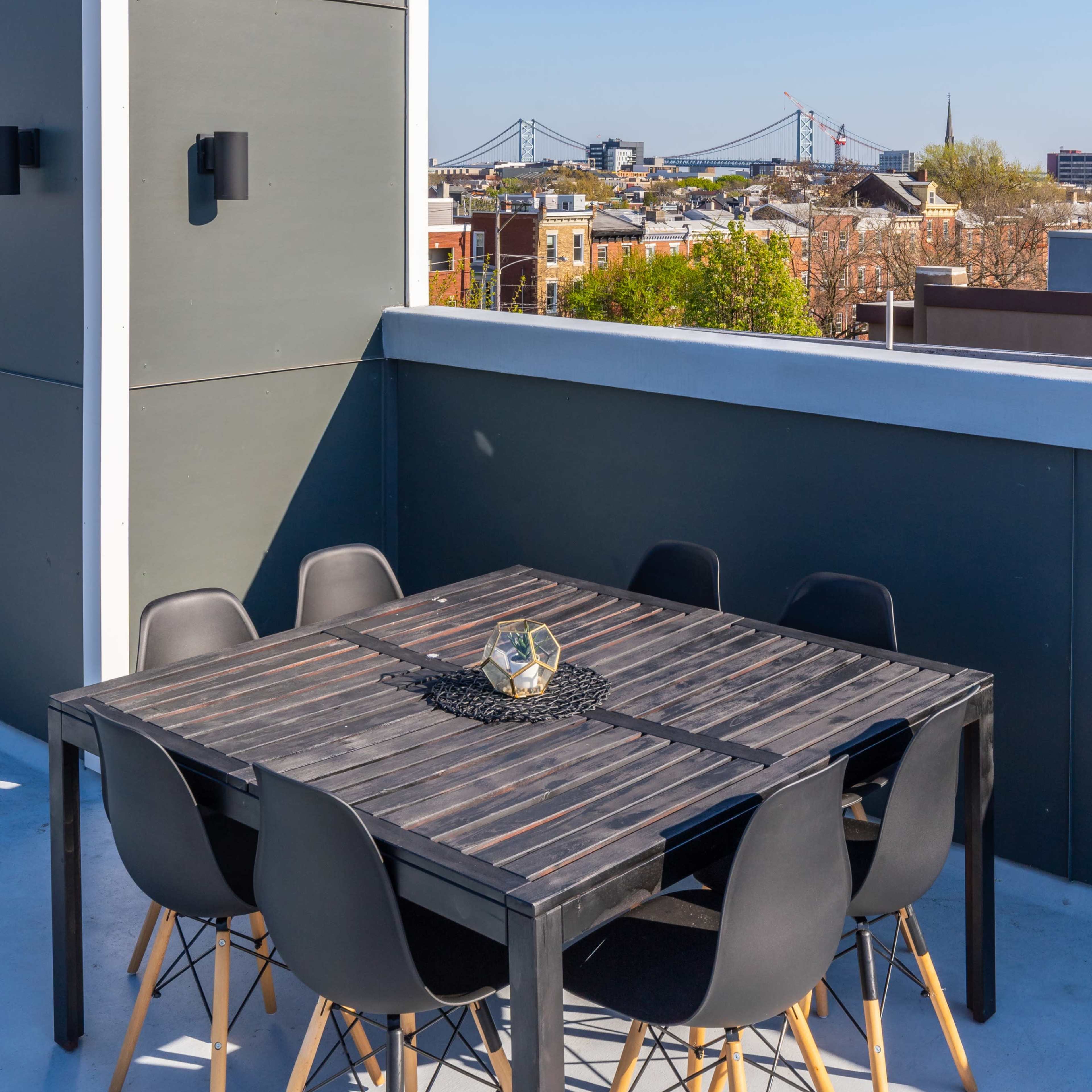 A rooftop terrace features a wooden table surrounded by black chairs, with a view of urban buildings and a bridge in the distance.