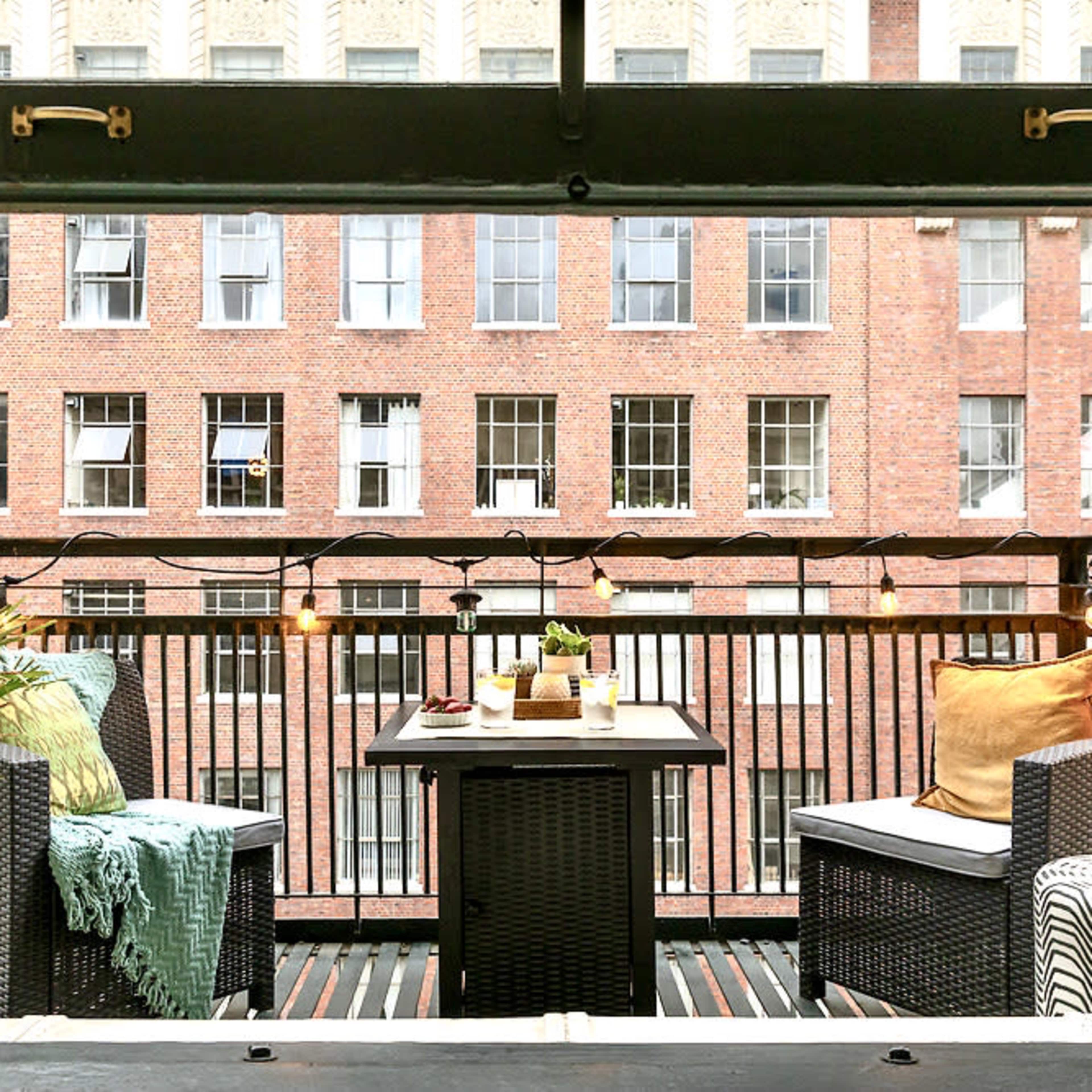 A balcony with two chairs and a table, surrounded by string lights and overlooking brick buildings.