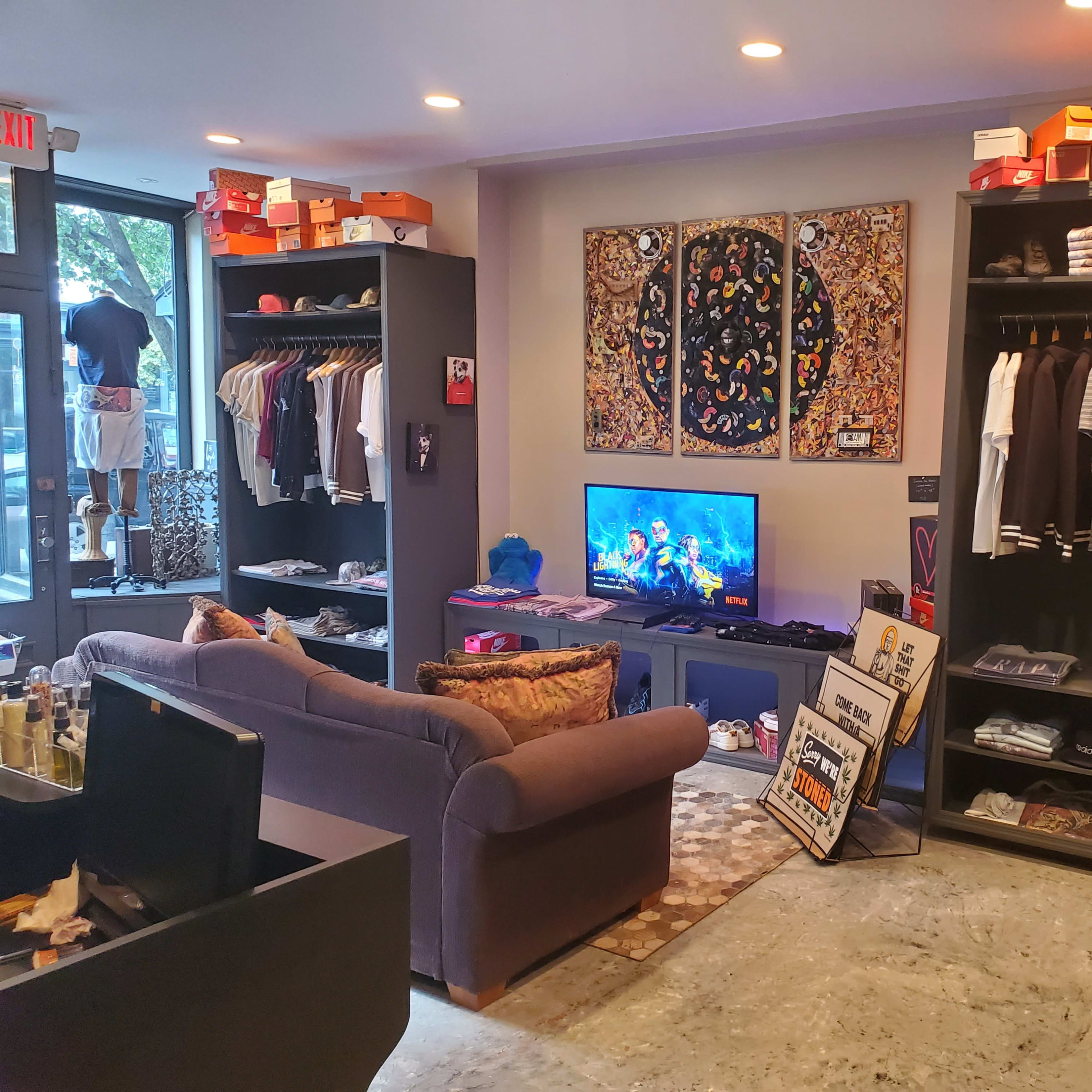 A clothing store interior featuring a sofa, racks of apparel, a television displaying colorful visuals, and various shoe boxes displayed on shelves.