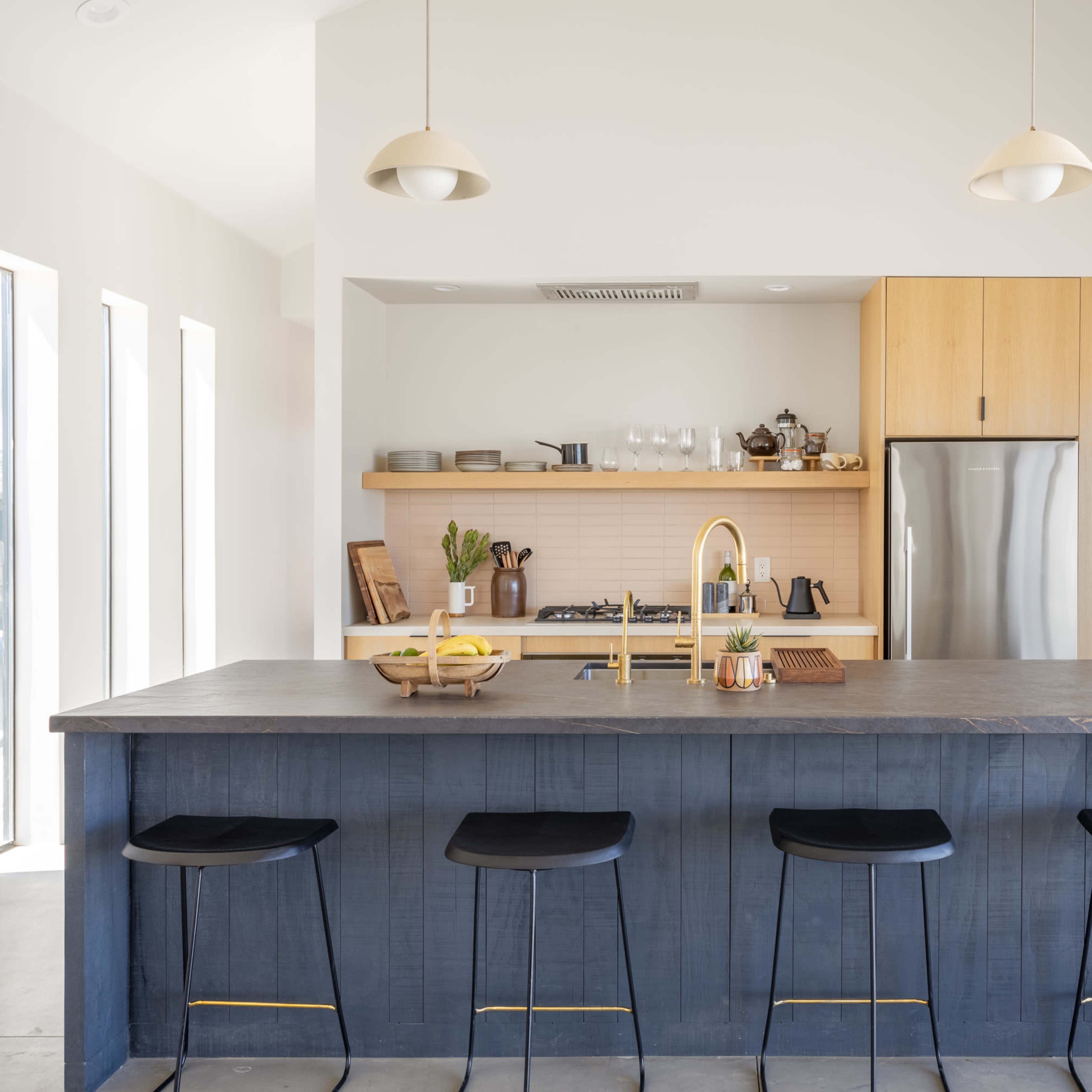 A modern kitchen featuring a large dark island with four bar stools, a stainless steel refrigerator, and light wood cabinetry.