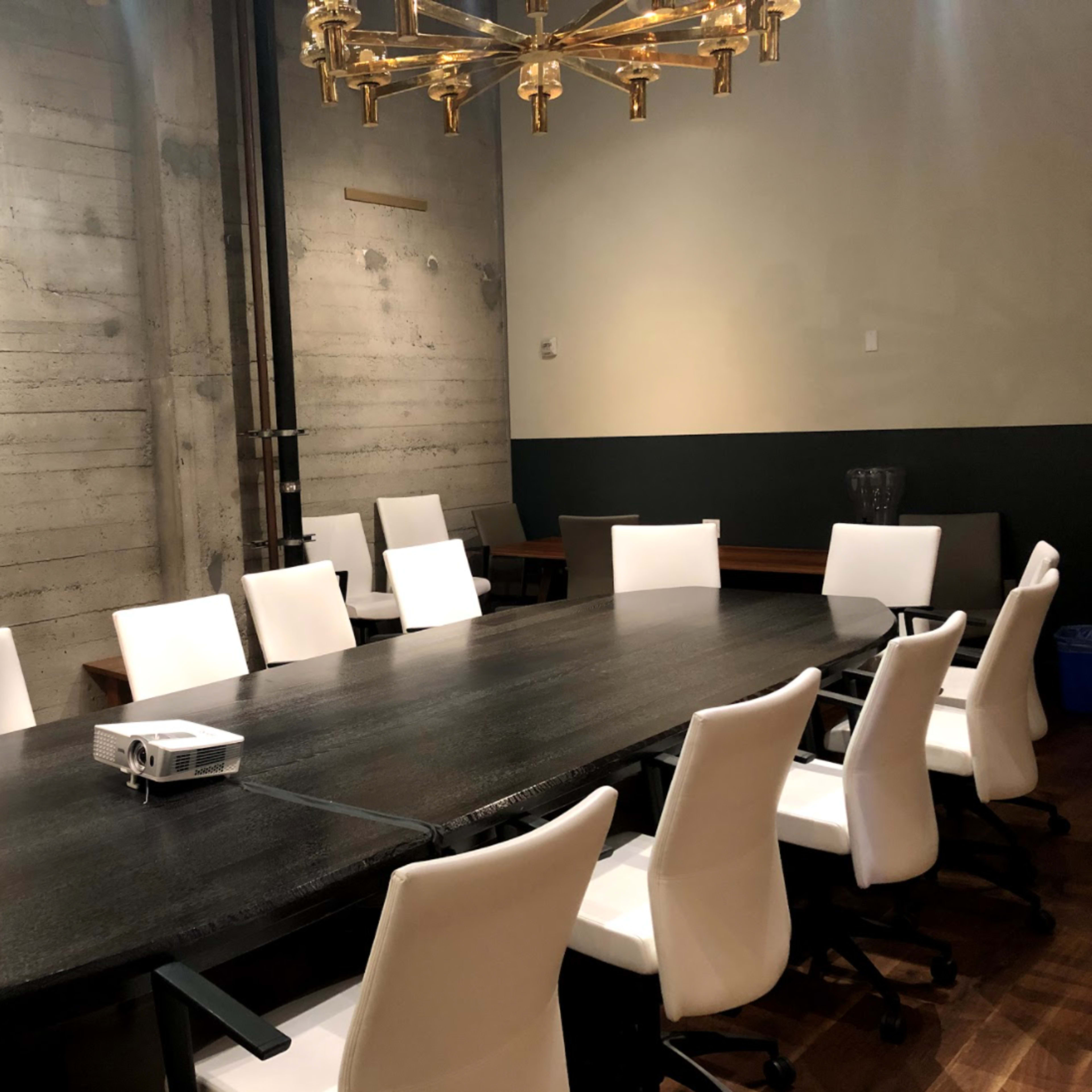 The room features a large, dark wooden conference table surrounded by white swivel chairs, with a projector placed at one end.