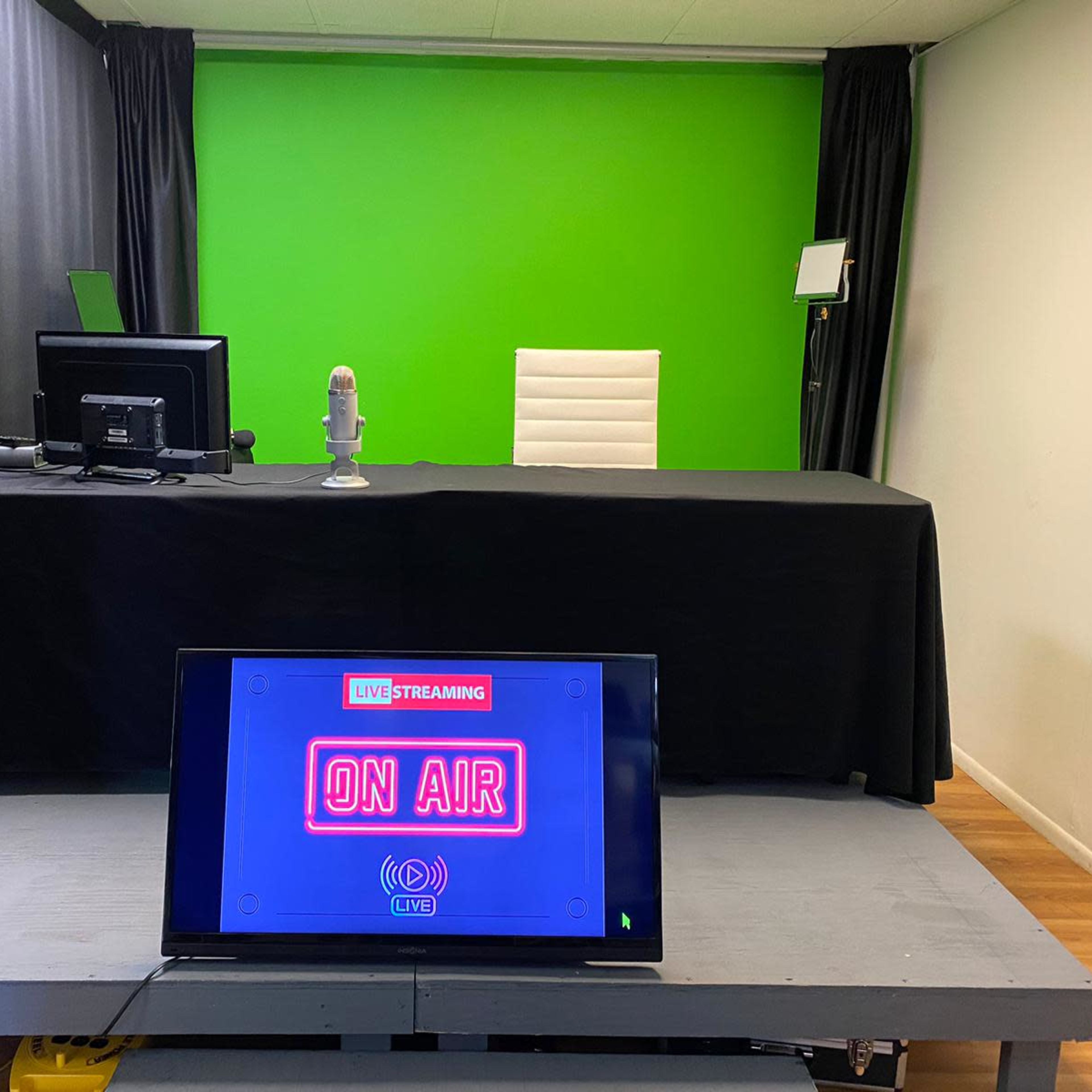 A streaming setup with a green screen, a microphone on a table, a desk with a computer, and a TV displaying "LIVE STREAMING ON AIR."