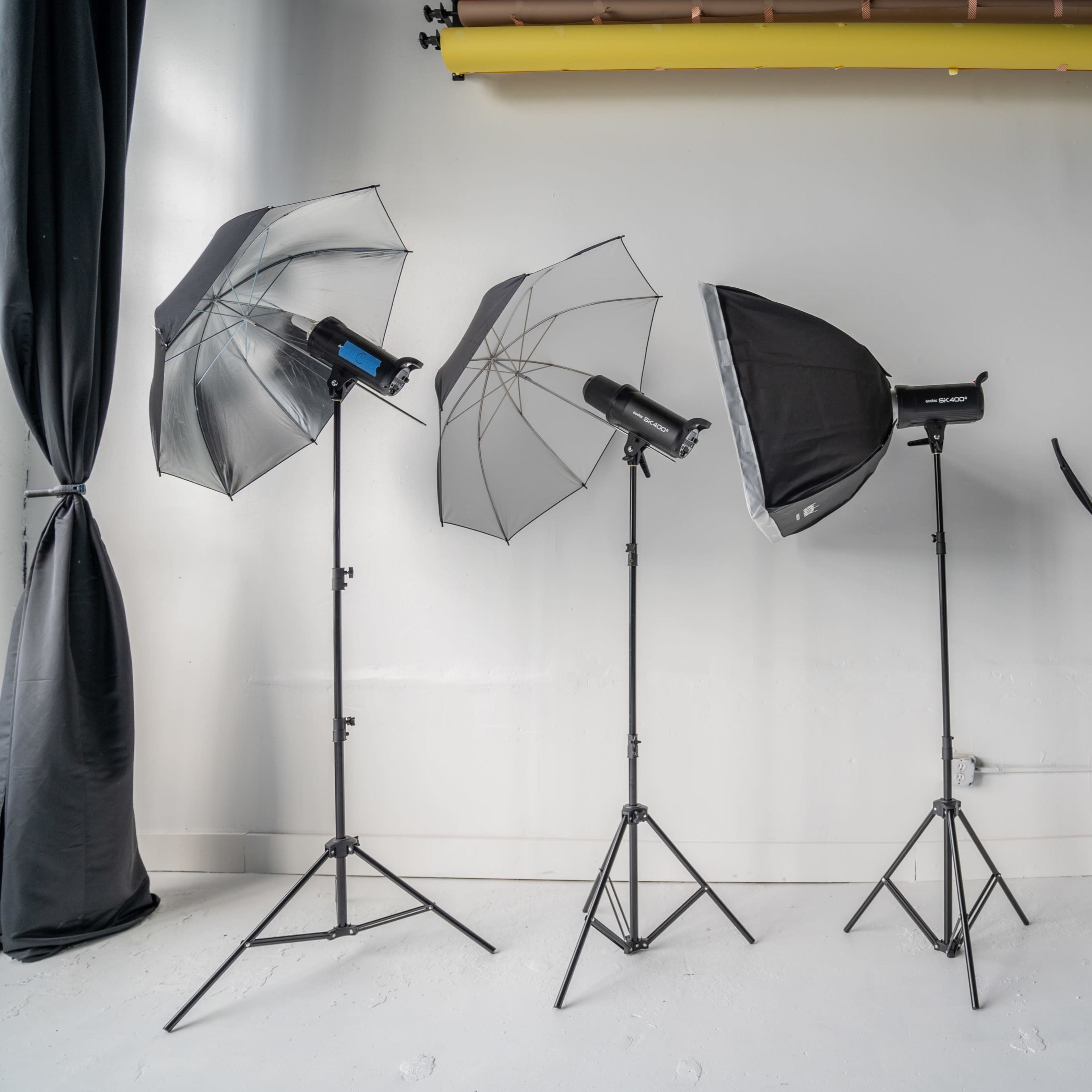 Four lighting setups on stands, including two umbrella lights and a softbox, in a studio with a gray wall and backdrop.