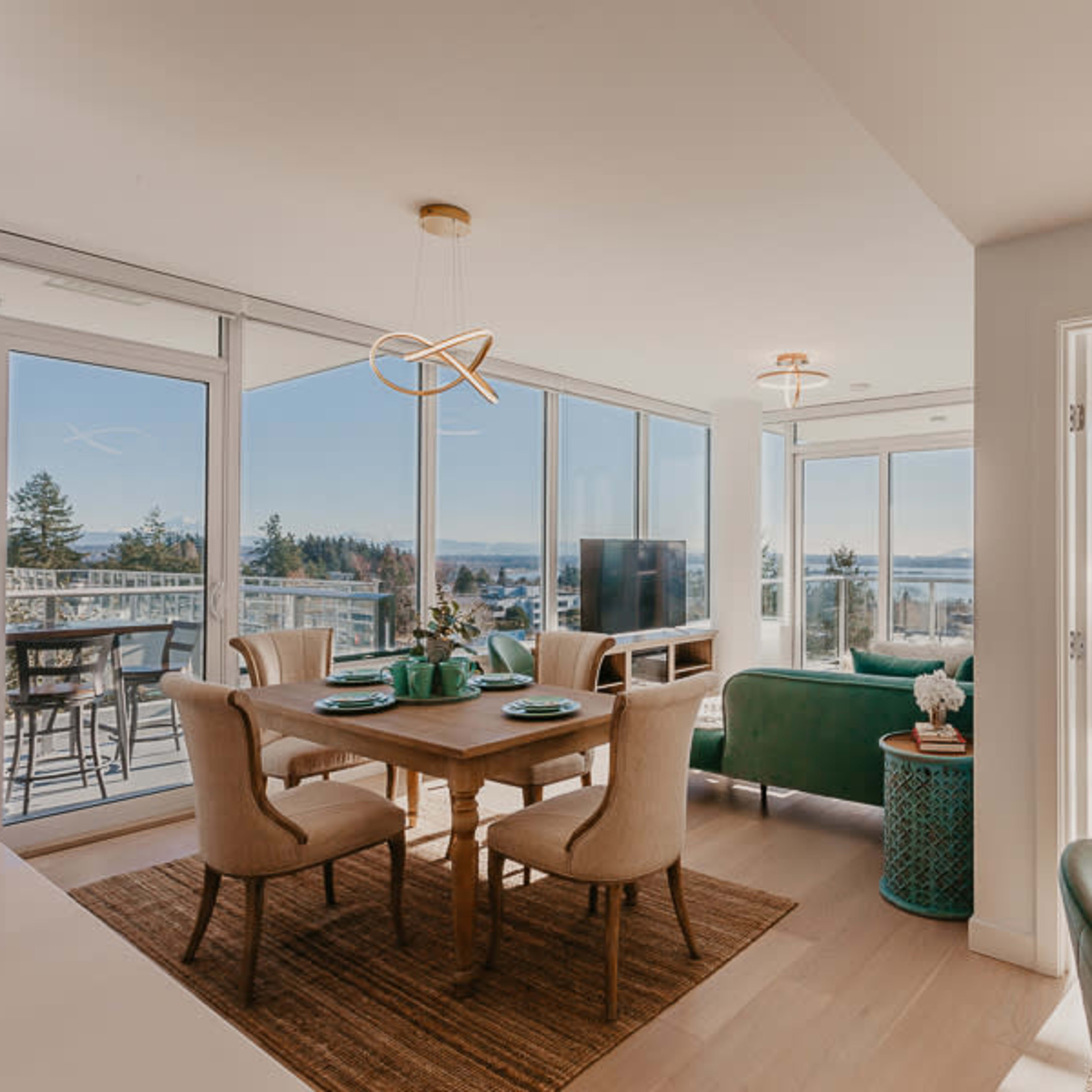 A modern open-concept living and dining area with large windows offering views of the outdoors, a dining table set for four, and a cozy seating area with a green couch.