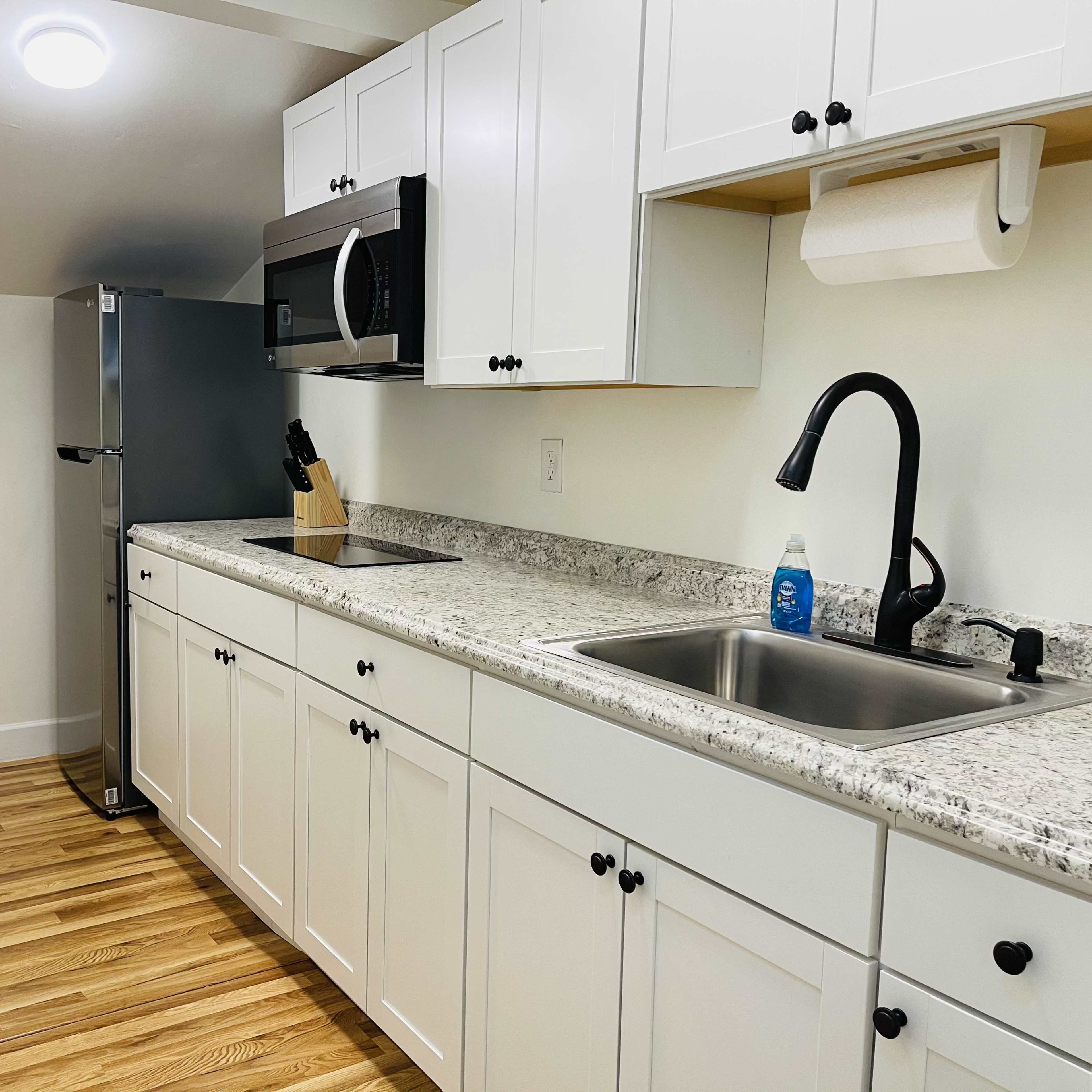 A modern kitchen with white cabinets, a stainless steel refrigerator, a microwave, a sink, and wooden flooring.