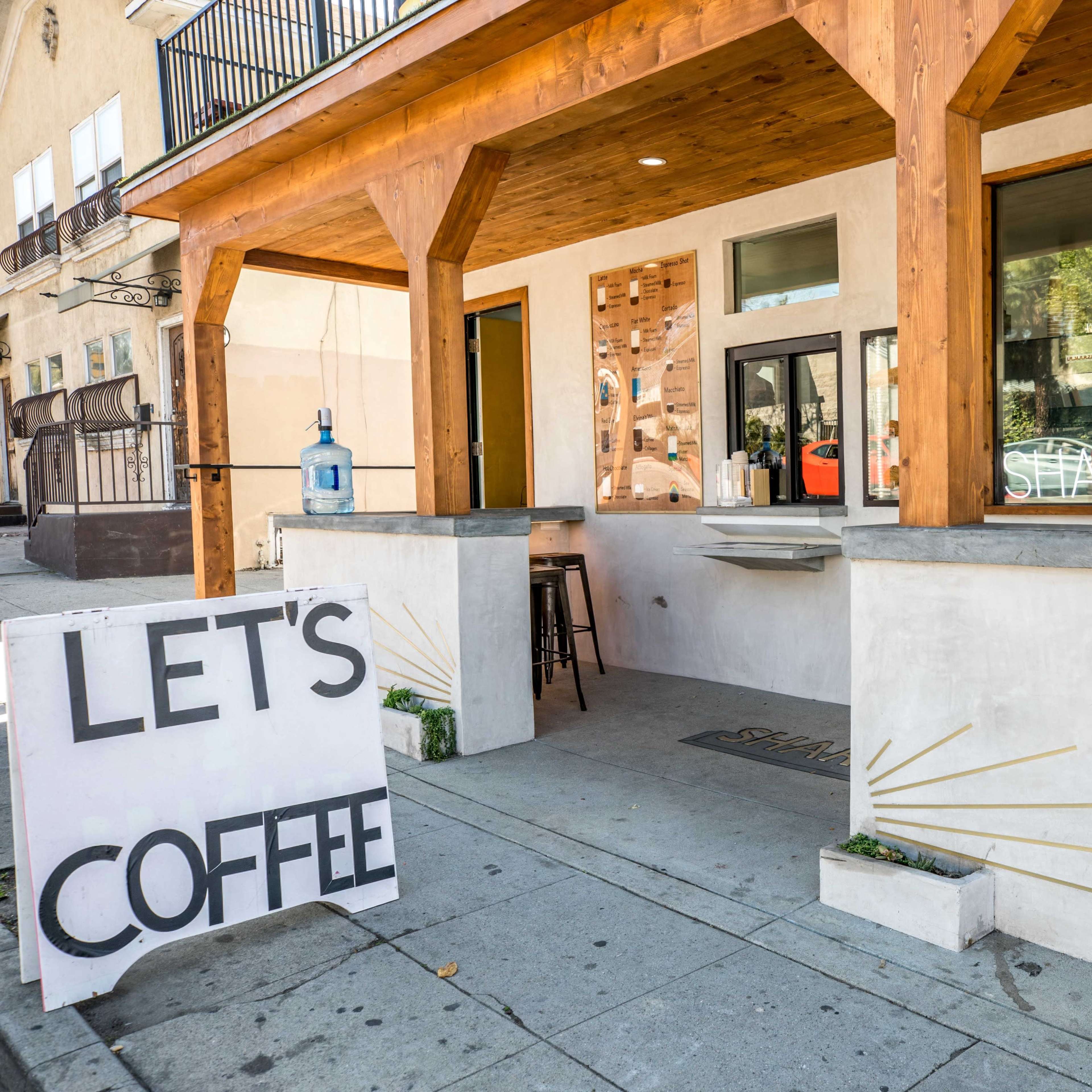 A coffee shop with a wooden overhang features an open window and a sign reading "LET'S COFFEE" outside.