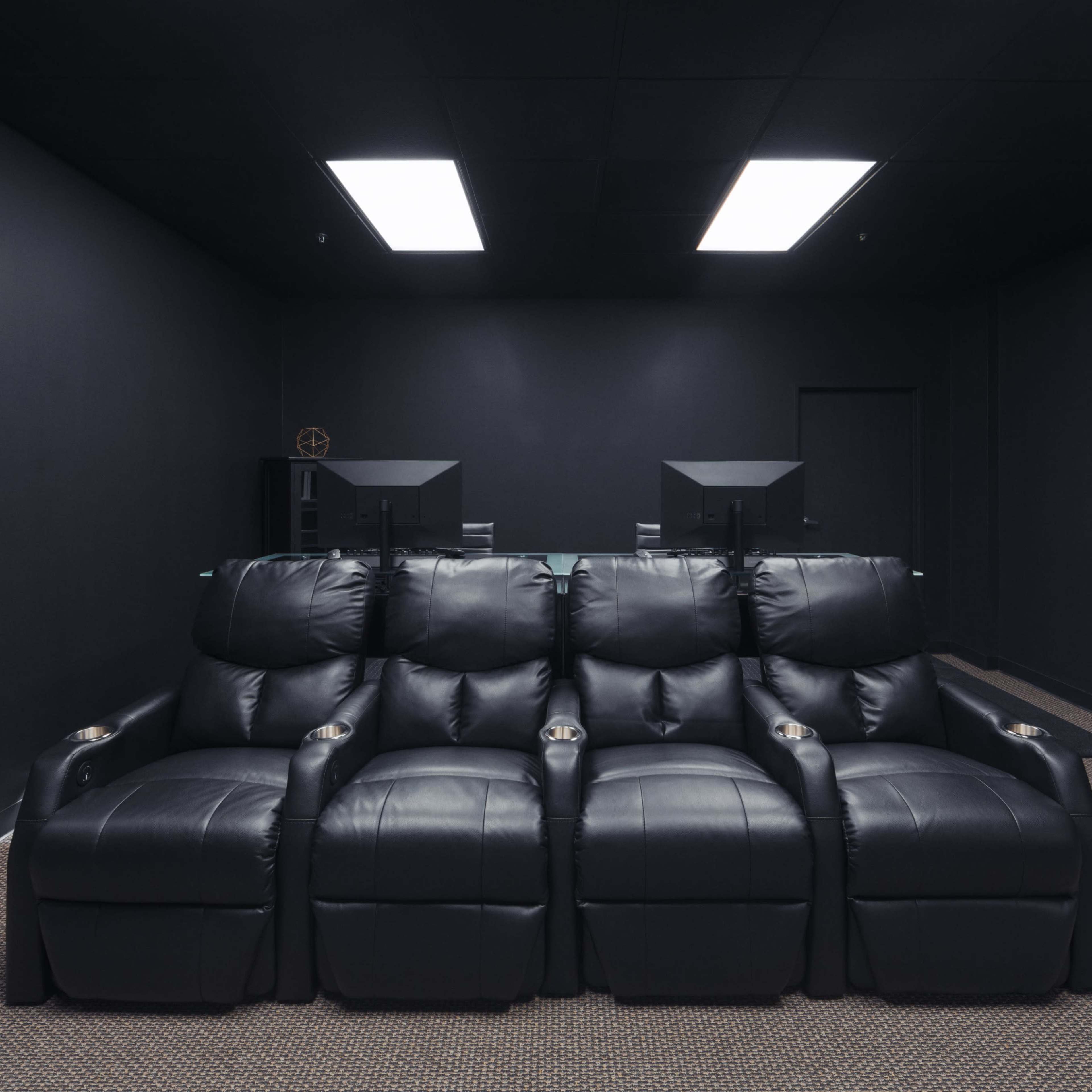 A dimly lit home theater room featuring a row of four black reclining seats with cup holders, positioned against a dark, minimalistic backdrop.