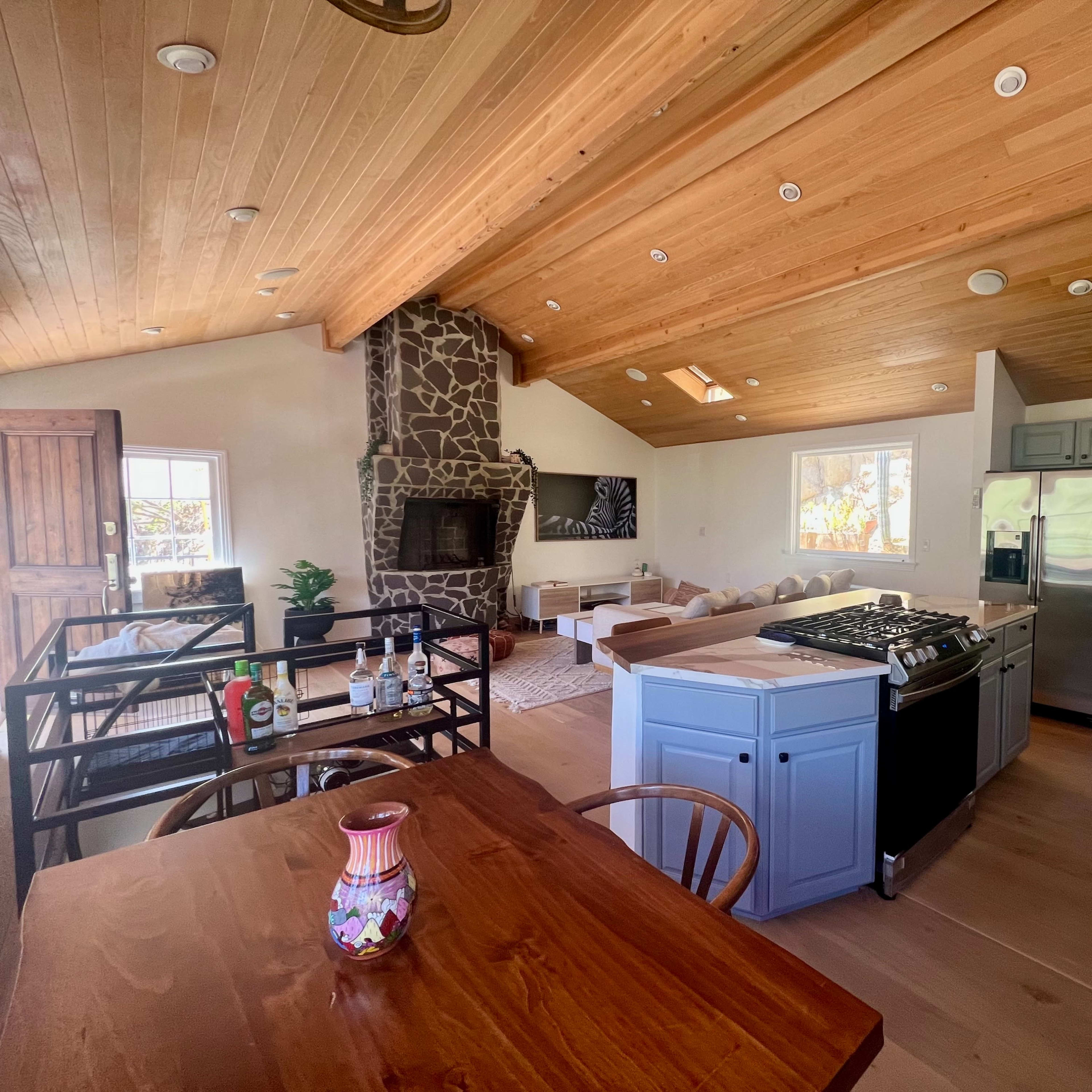 An open-concept living space featuring a wood-beamed ceiling, a stone fireplace, a kitchen with blue cabinetry and stainless steel appliances, and a dining area with a wooden table.
