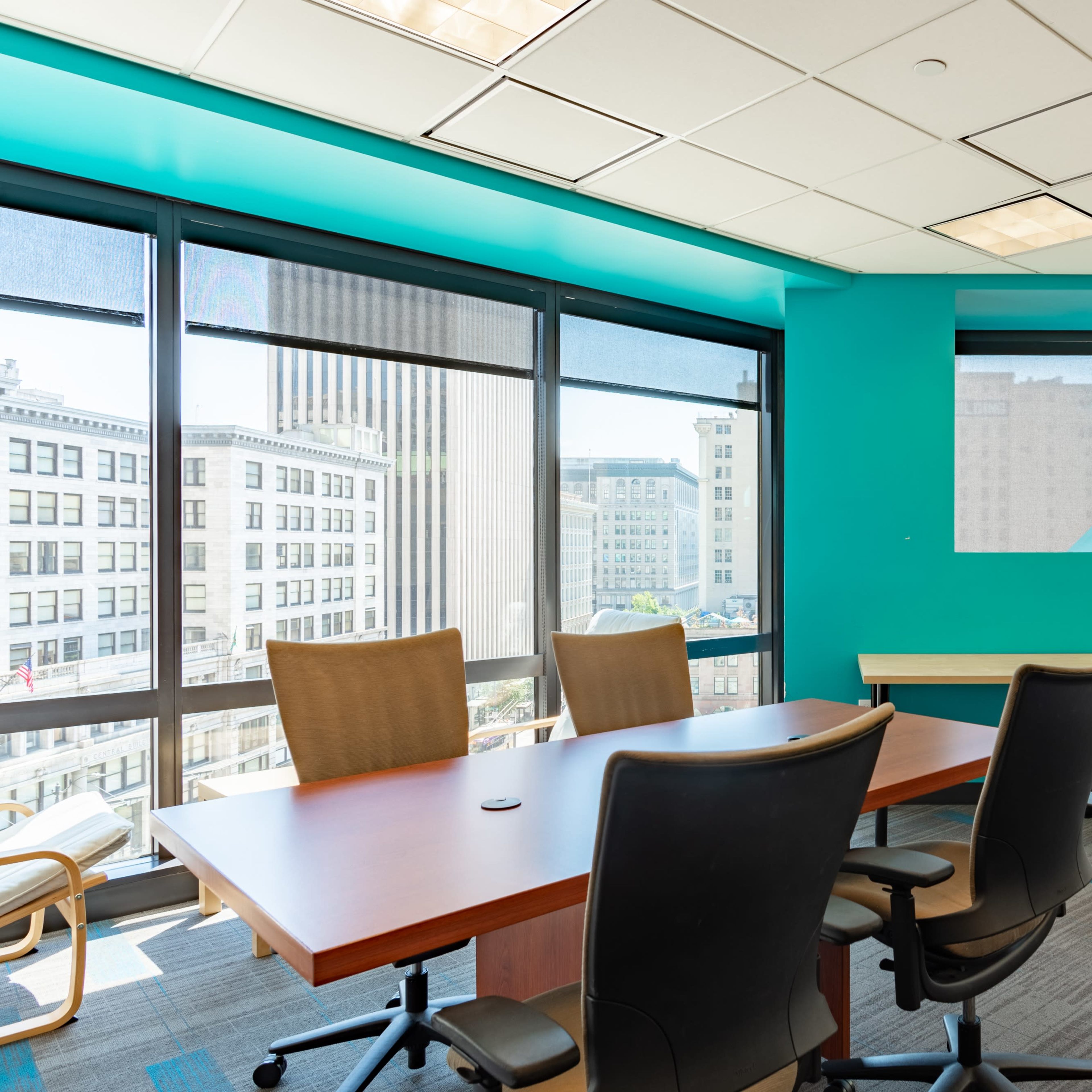 A modern conference room features a large wooden table surrounded by office chairs and large windows with a view of a cityscape.