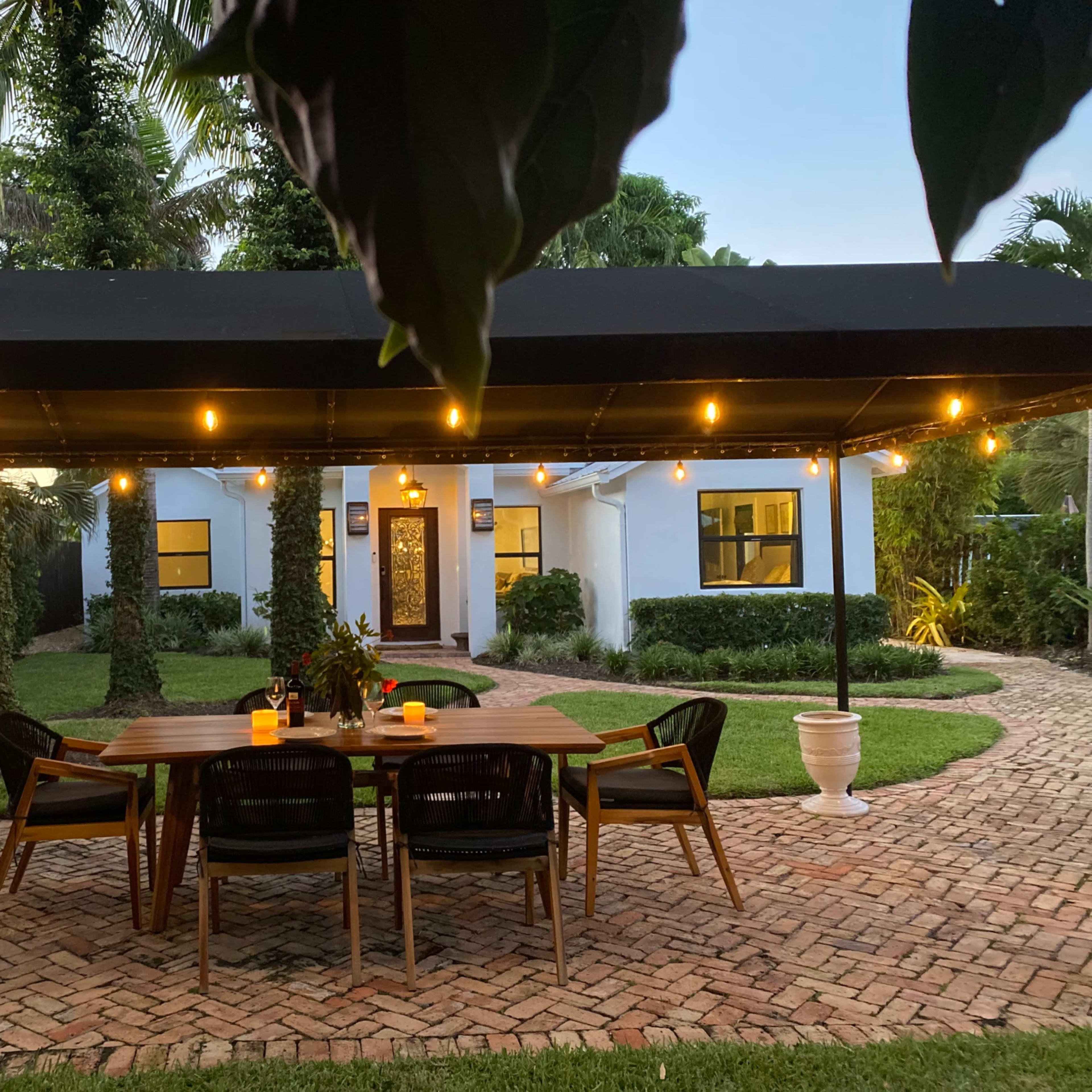 A patio with a wooden table and chairs is illuminated by string lights, leading to a house surrounded by greenery.