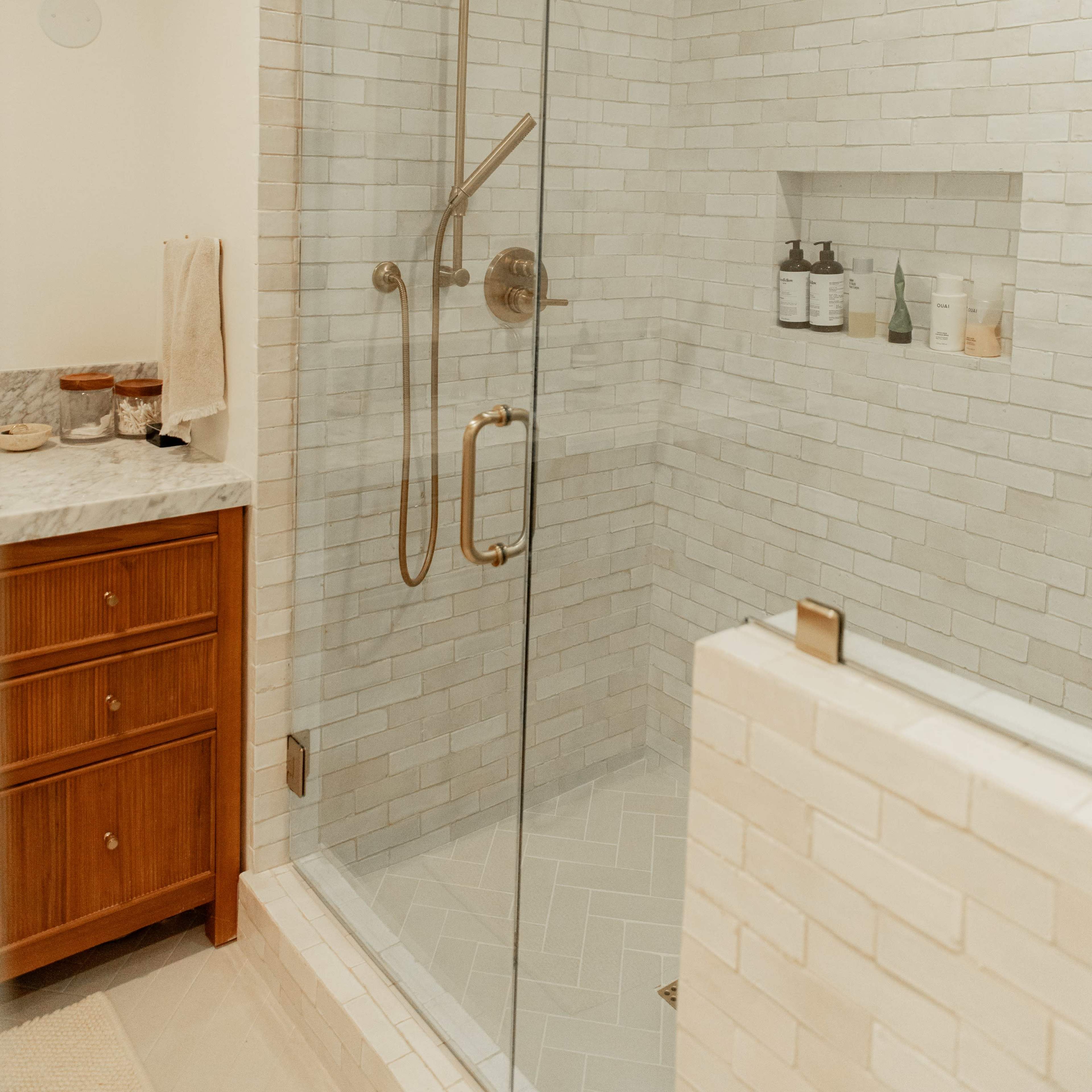 A modern bathroom featuring a glass shower stall with a rainfall showerhead and white tiled walls, alongside a wooden vanity with various toiletries.