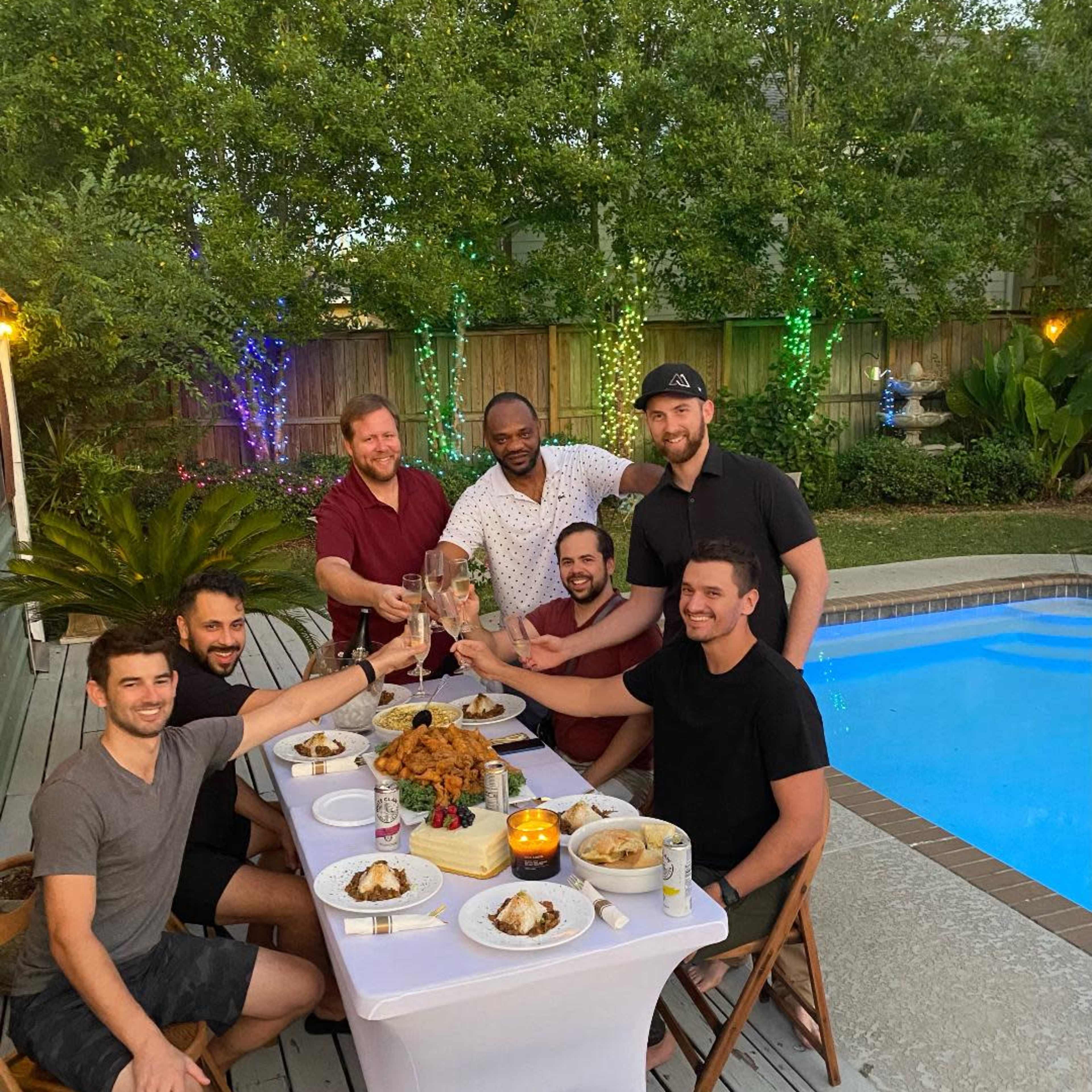 A group of seven men toast with drinks at a table set near a pool, surrounded by greenery and evening lights.