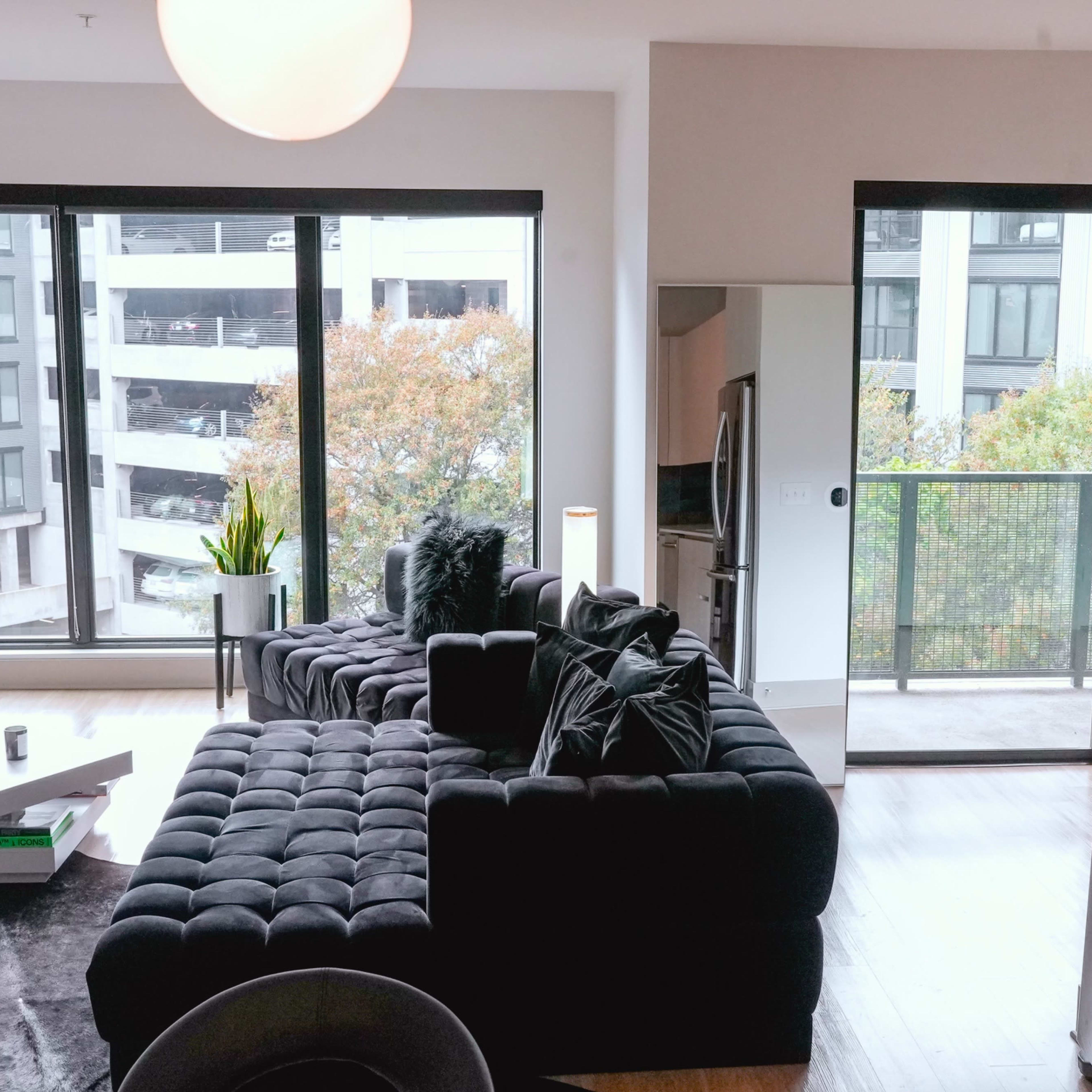 A modern living room features a black sectional sofa, large windows overlooking a balcony, and sleek kitchen appliances.