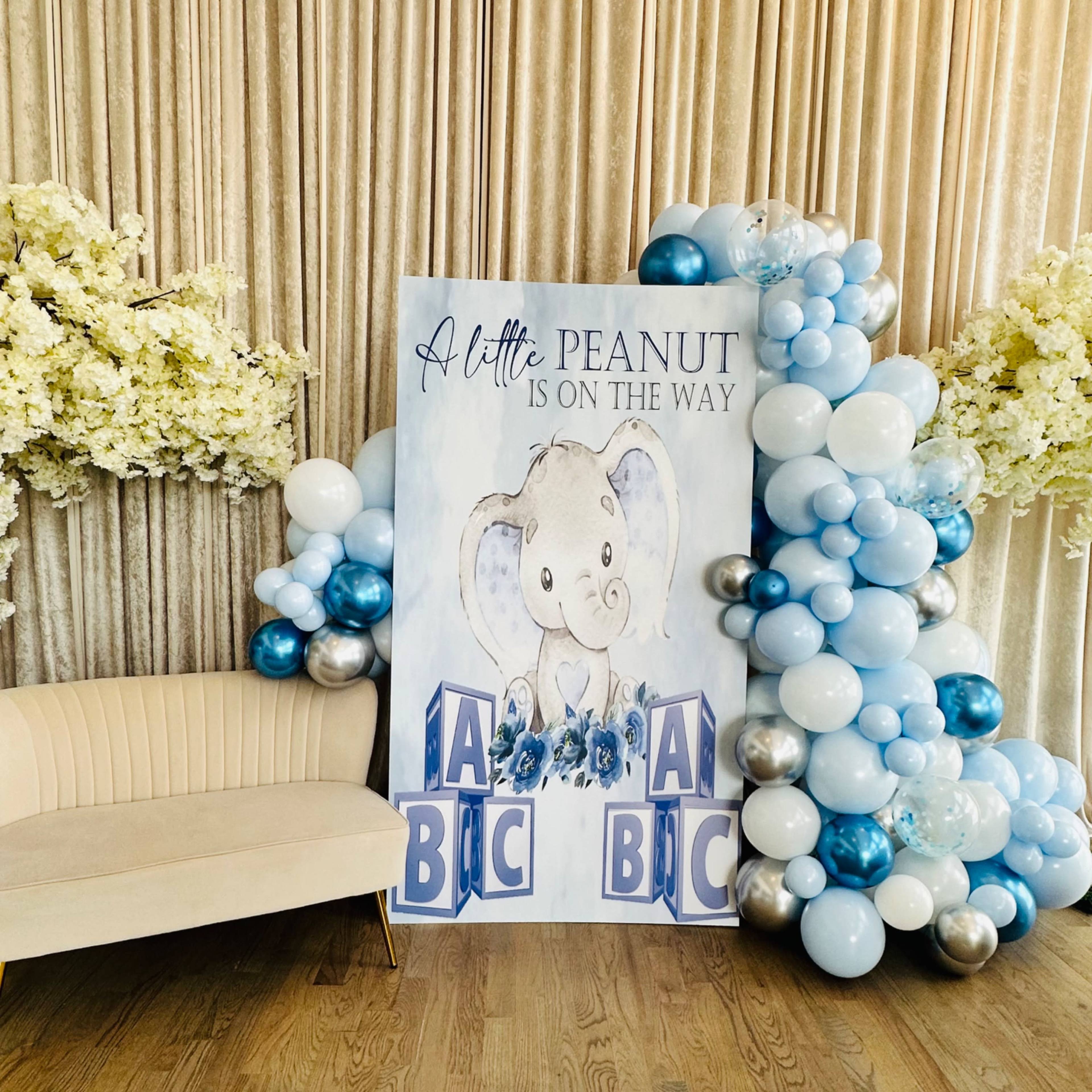 A decorative setup featuring a large sign with an elephant and the text "A little peanut is on the way," surrounded by clusters of blue and silver balloons and floral arrangements, with a cream-colored sofa in front.
