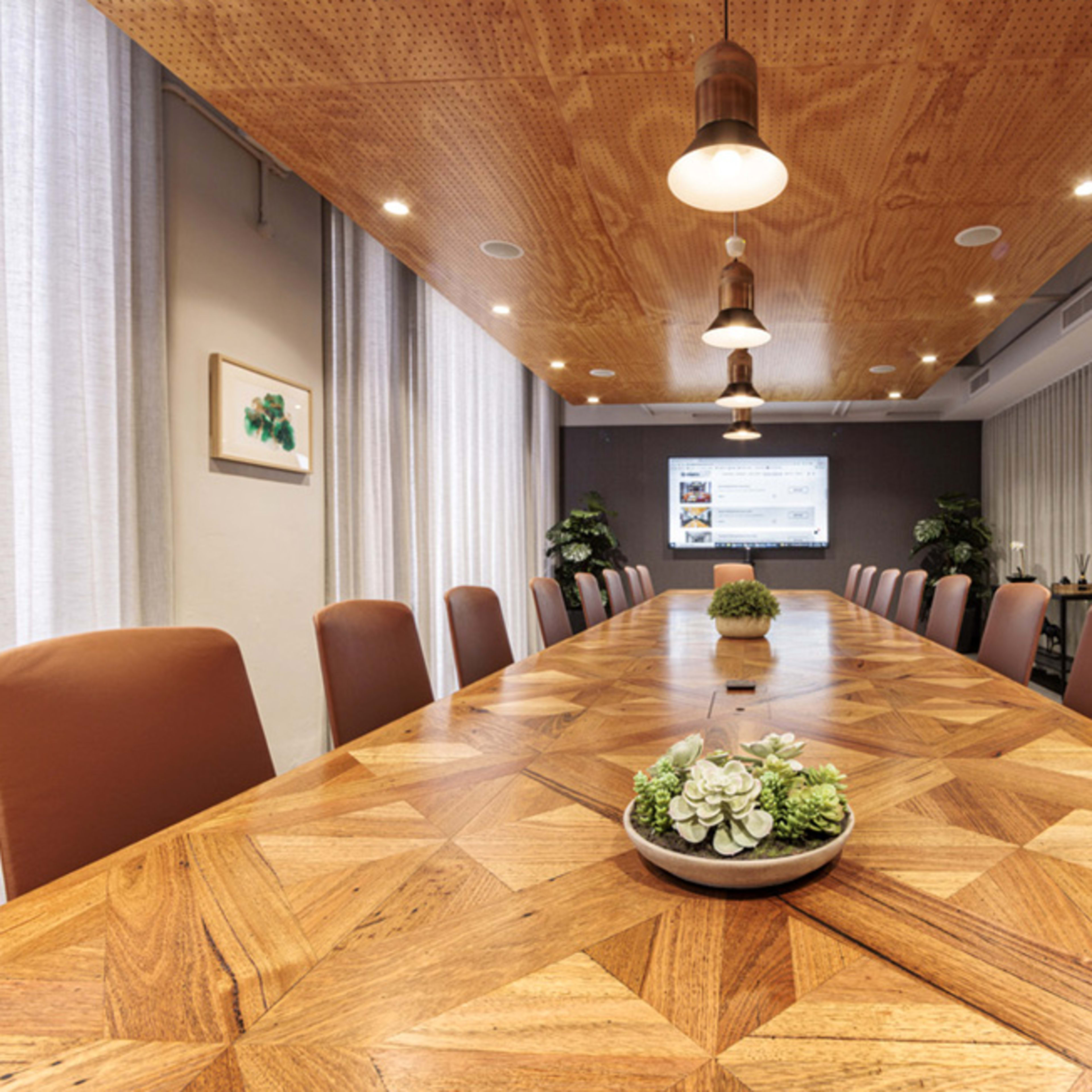 A large conference room features a wooden table with a geometric design, surrounded by brown chairs, and is illuminated by hanging lights with a screen displaying content at one end.