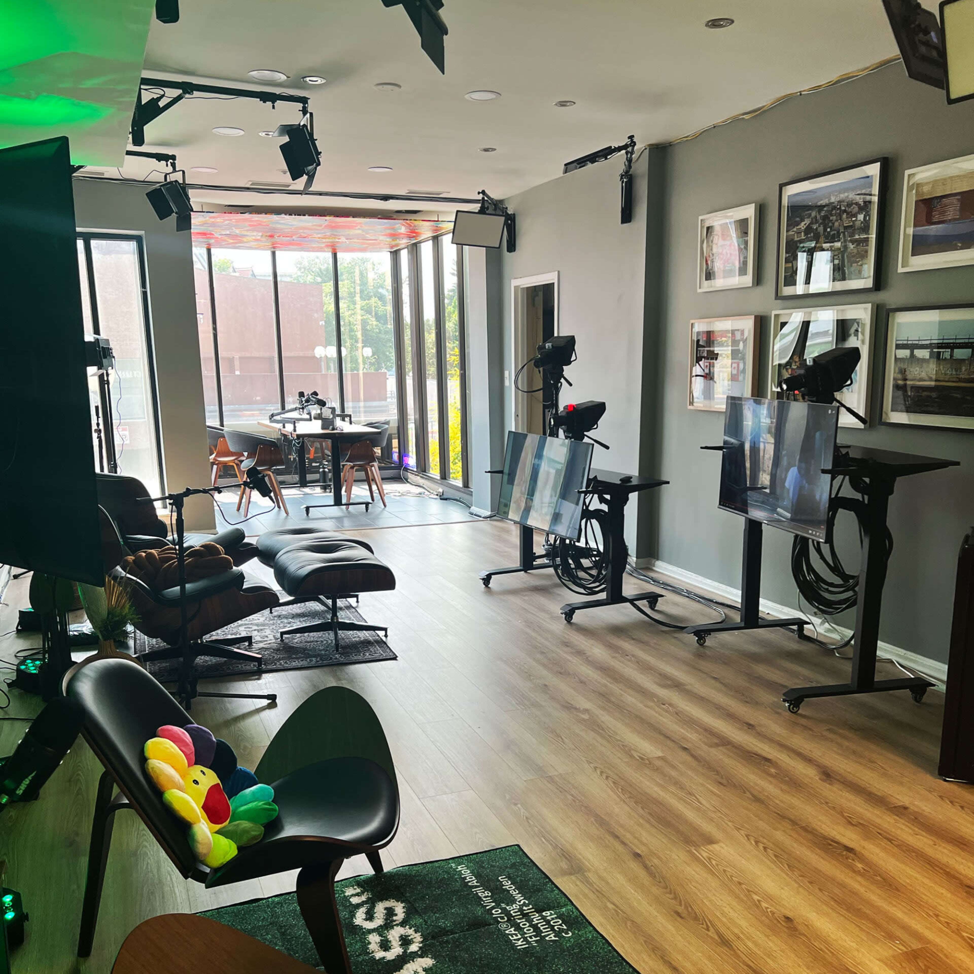 A modern studio with several cameras, chairs, and screens arranged in a well-lit space featuring large windows and wooden flooring.