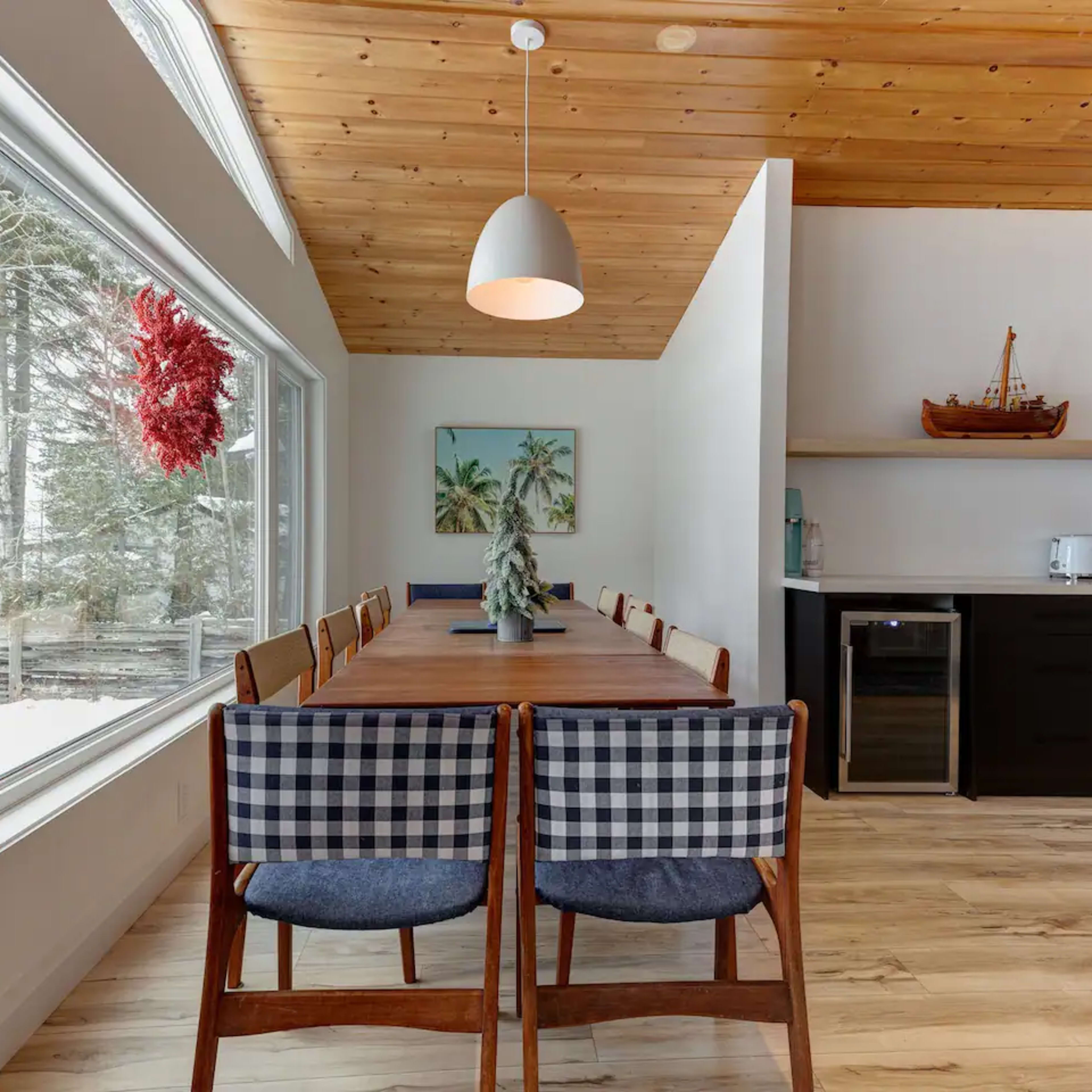 A long wooden dining table with checkered chairs is positioned next to large windows that overlook a snowy landscape, while a modern kitchen area is situated in the background.