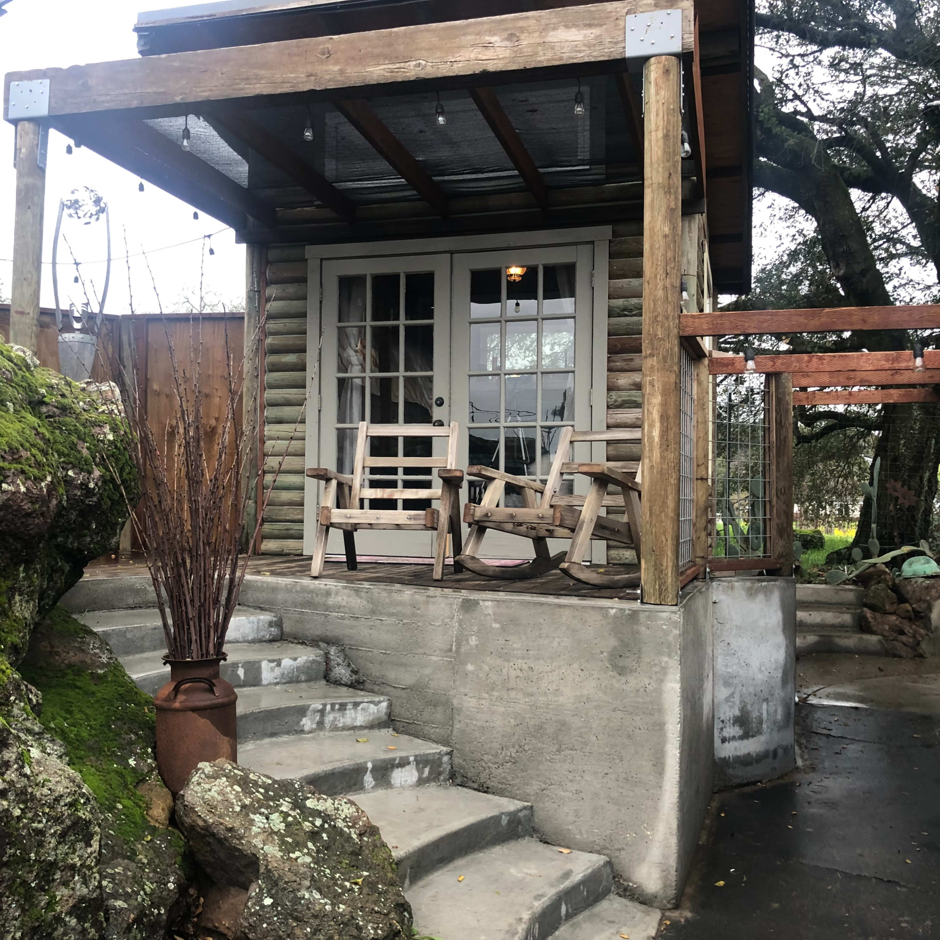 A small wooden cabin with a covered porch and two chairs, surrounded by large rocks and greenery.