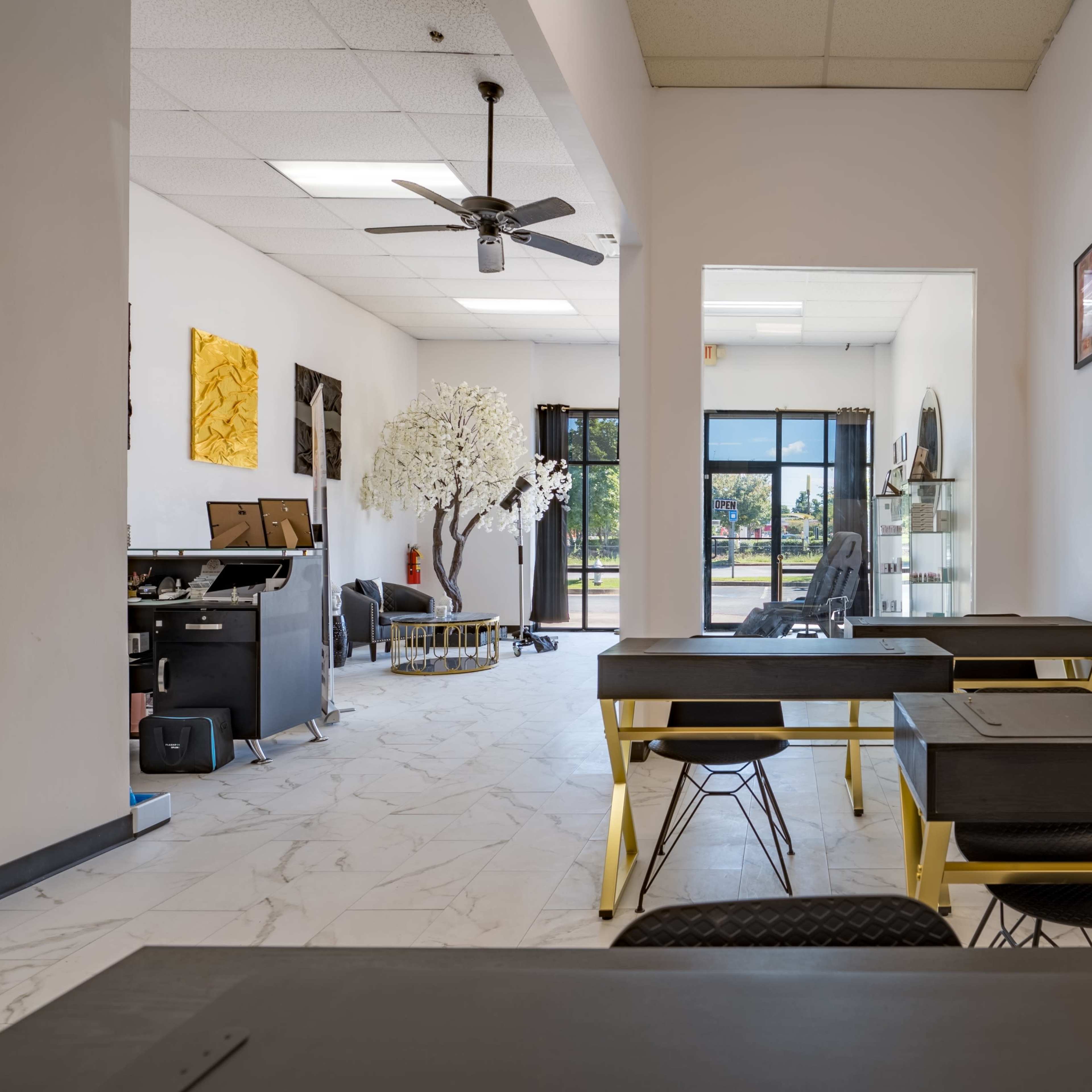 A modern workspace featuring several gray desks with gold legs, a large open area, and a decorative tree, with glass doors leading to an outdoor view.